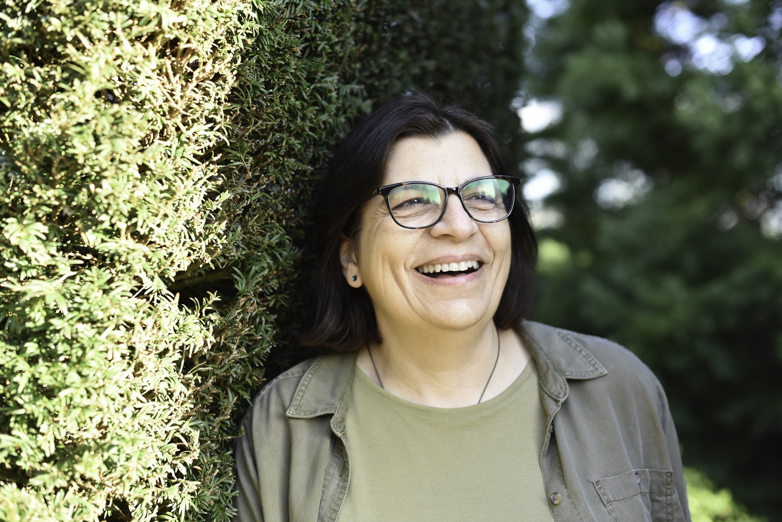 Anna Zannides — British author of How Did I Get Here? — smiling, with a natural green background.