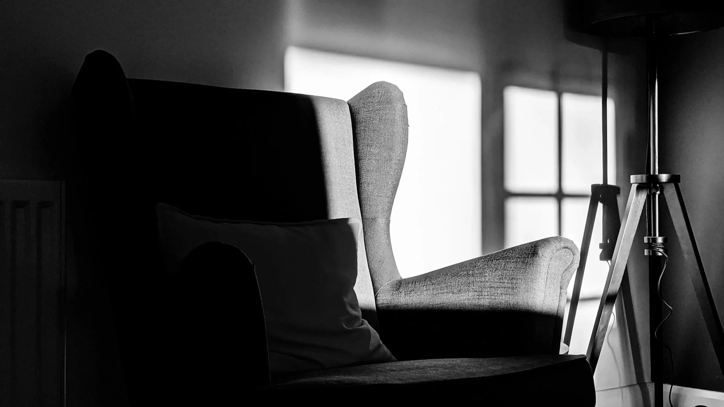 A black and white photograph of an empty armchair in a dark room, with a ray of light coming in from behind.
