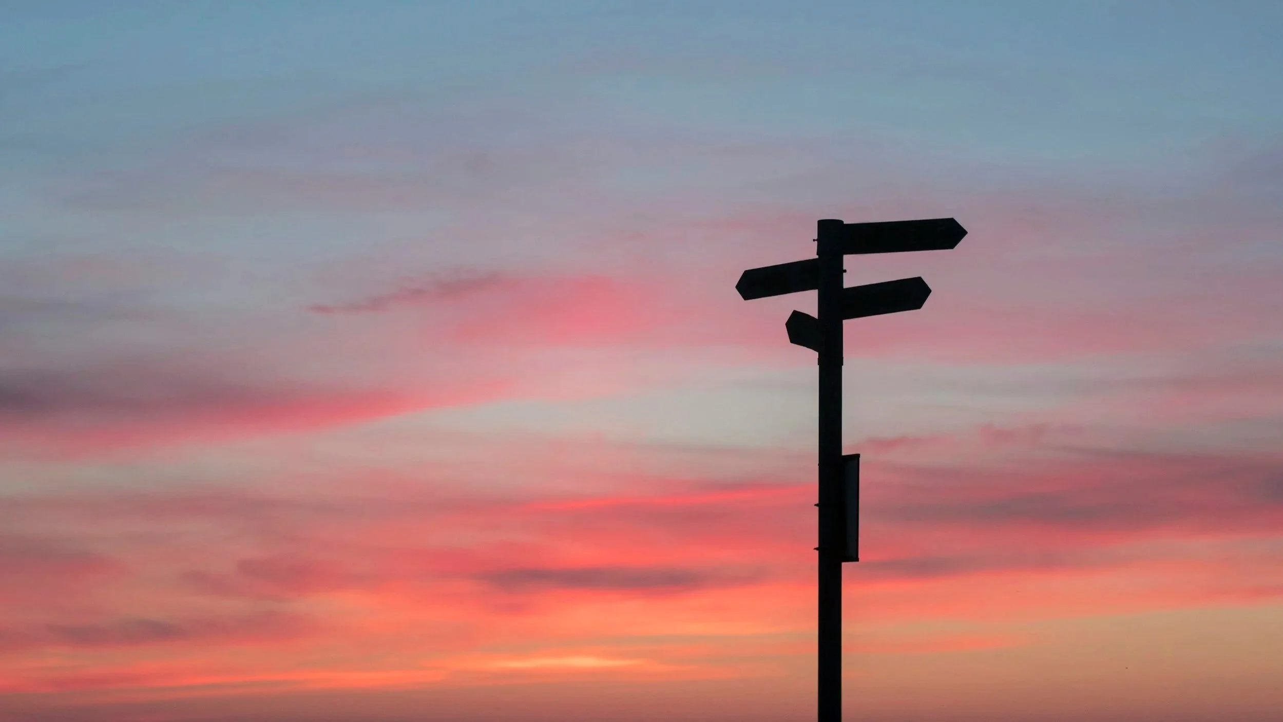 Wooden signpost pointing in multiple directions against a soft pink evening sky.