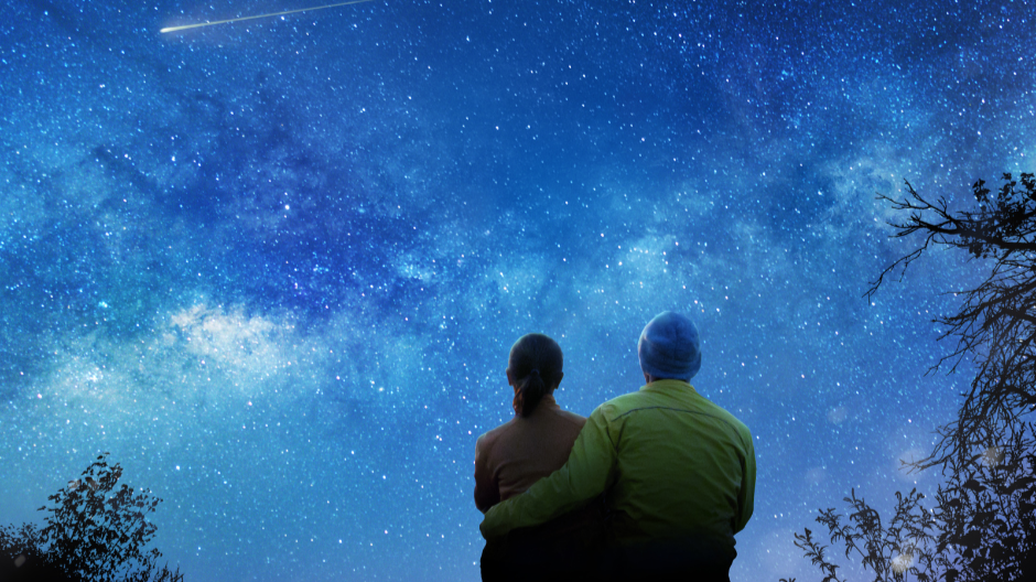 Couple sitting on a bench under a starry night sky, looking upward in quiet reflection.