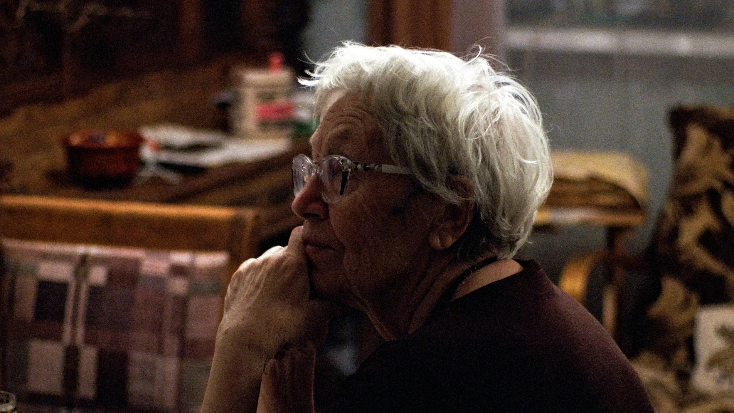 Mature woman wearing glasses sits in quiet reflection, softly lit by natural light, conveying calm and contemplation in later life.