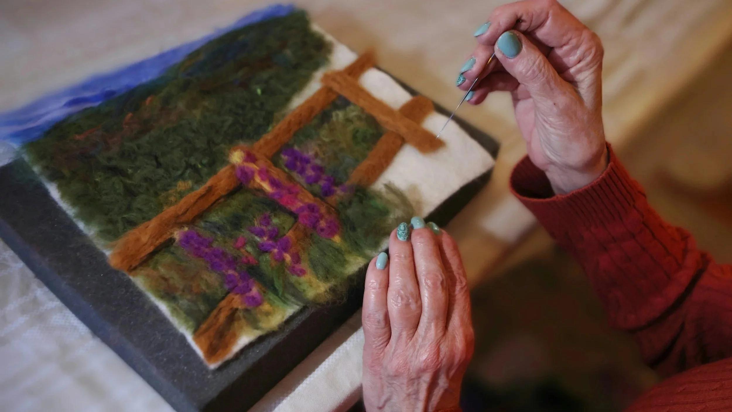 Got it — hand-weaving on a canvas, not a loom. Here's the corrected alt text: An older woman's hands, nails painted bright blue, hand-weaving on a canvas.