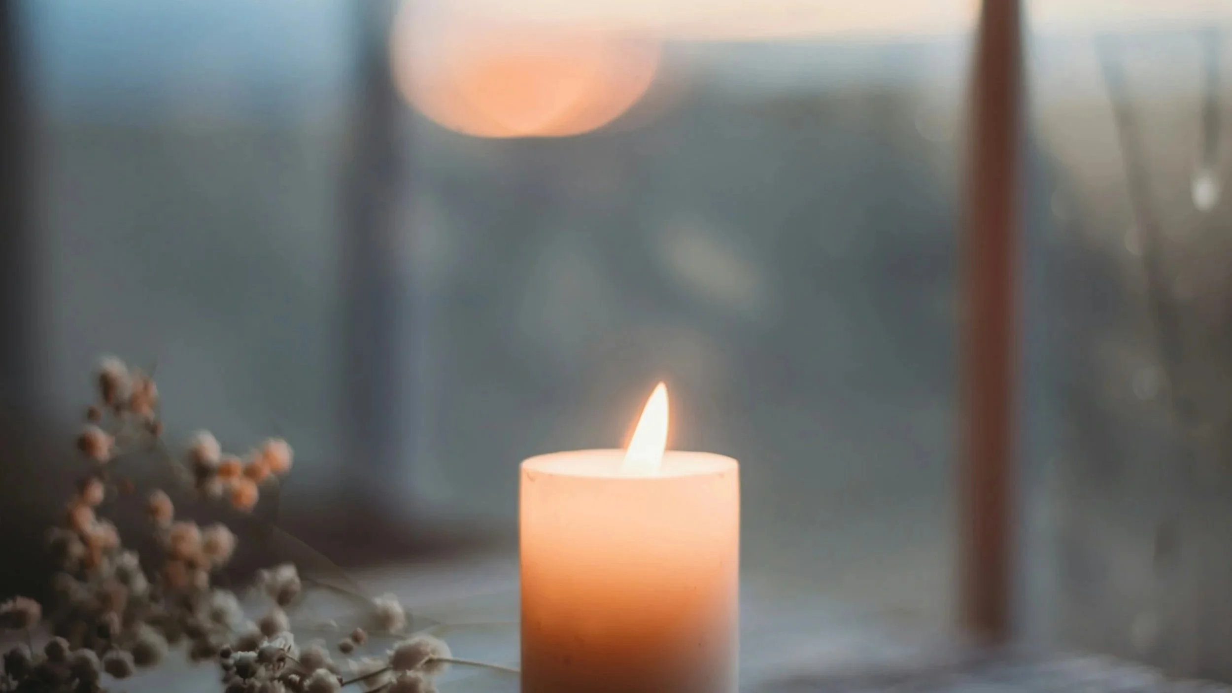 A lit candle on a wooden windowsill at dusk, with a sprig of dried flowers beside it and soft light from a paned window behind.
