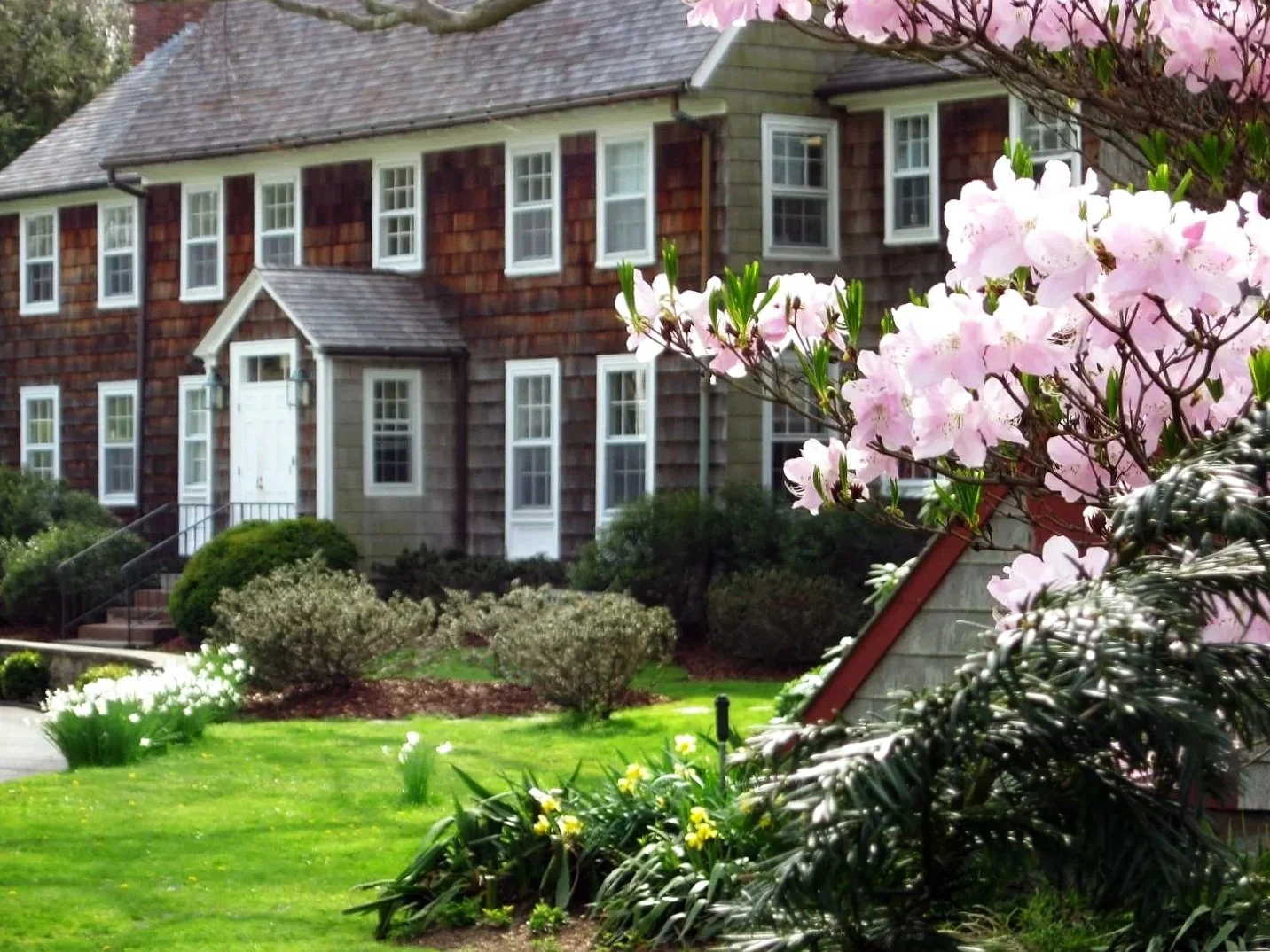 A two-story house with wooden shingles and multiple white-framed windows, surrounded by a green lawn and blooming pink and white flowers in the foreground during daytime.