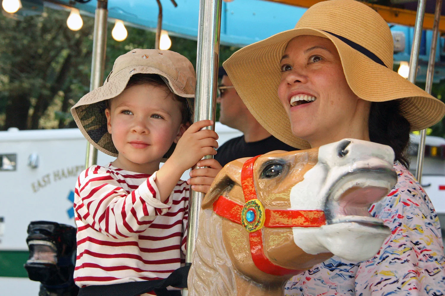 A woman and a young boy enjoying a carousel ride, with the woman smiling and the boy holding the carousel pole, both wearing wide-brimmed hats, nearby trees, lights, and a 'East Hummel' sign in the background.