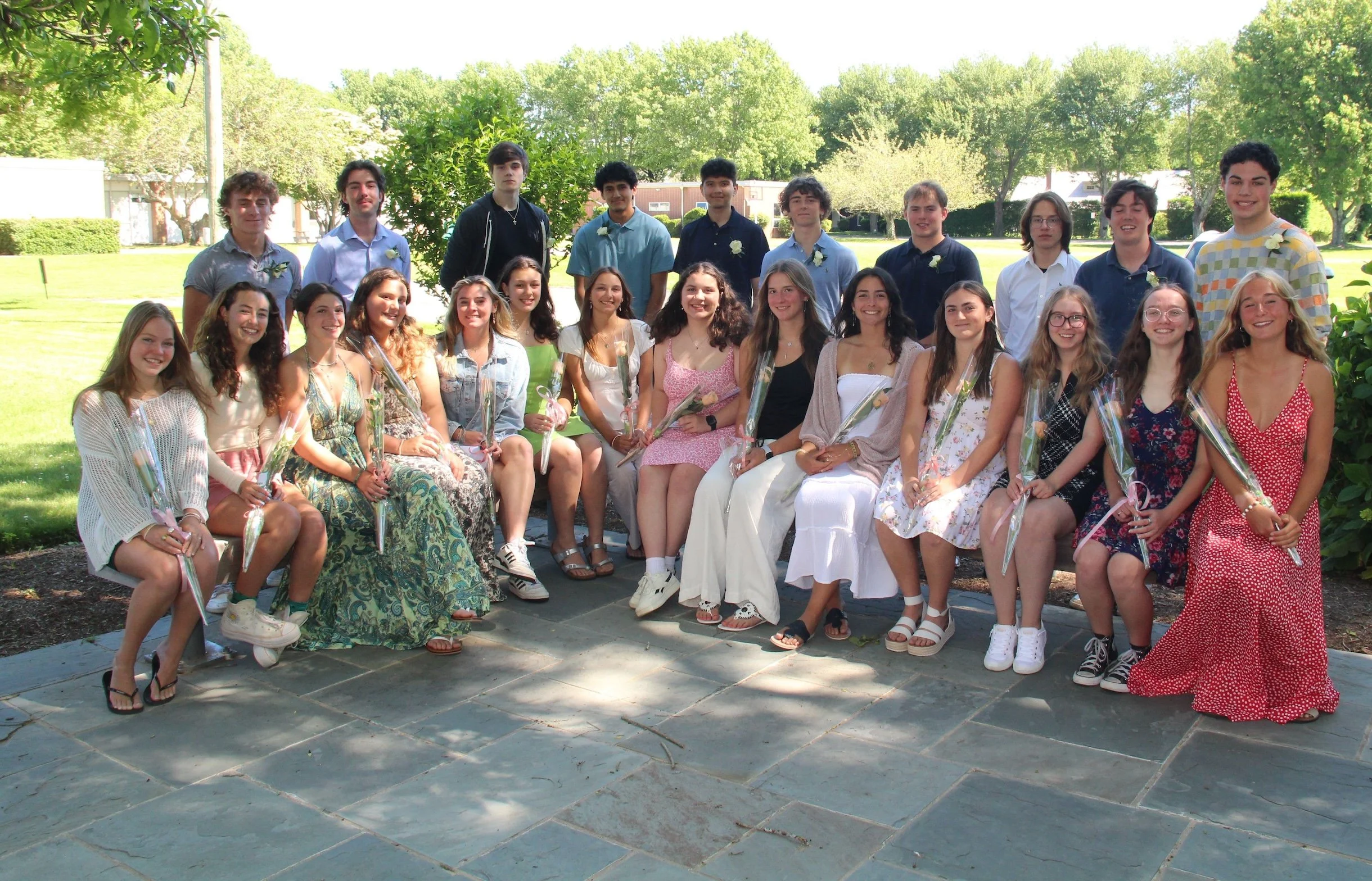 A group of young people, mostly teenagers, gathered outdoors on a sunny day. They are arranged in two rows, with some seated and others standing behind them. Many are holding flowers wrapped in plastic. The background features green trees and open grassy areas, suggesting a park or school yard setting.