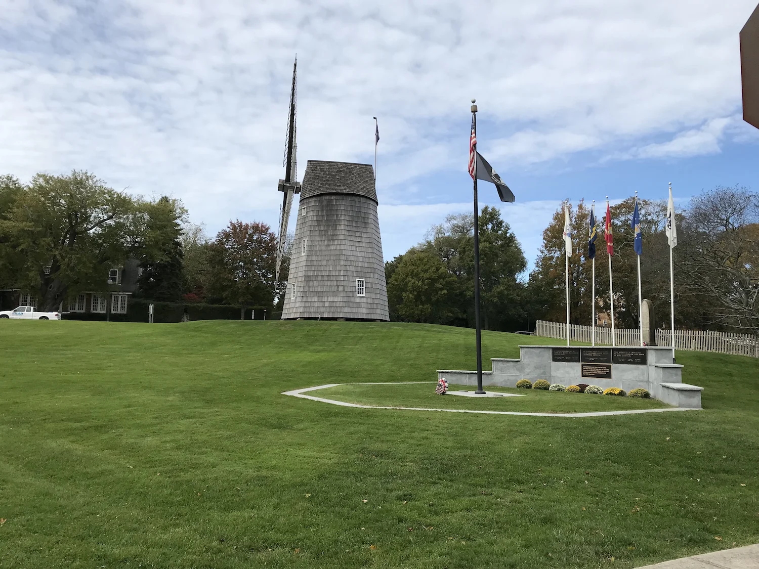 A memorial with several flags on poles, including the American flag, set on a grassy area with a concrete platform, and a windmill structure stands in the background among trees under a partly cloudy sky.