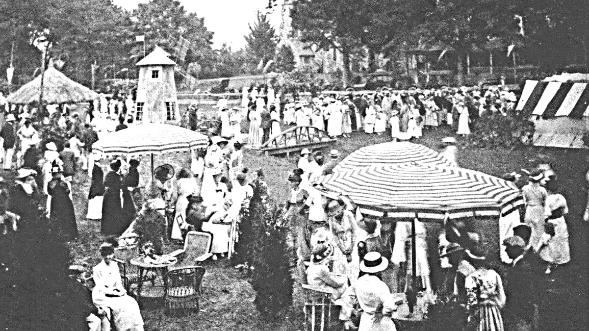 Black and white photo of a large outdoor gathering of people, many women in dresses and hats, some children, around tables, umbrellas, and a small tower, in a park setting with trees.