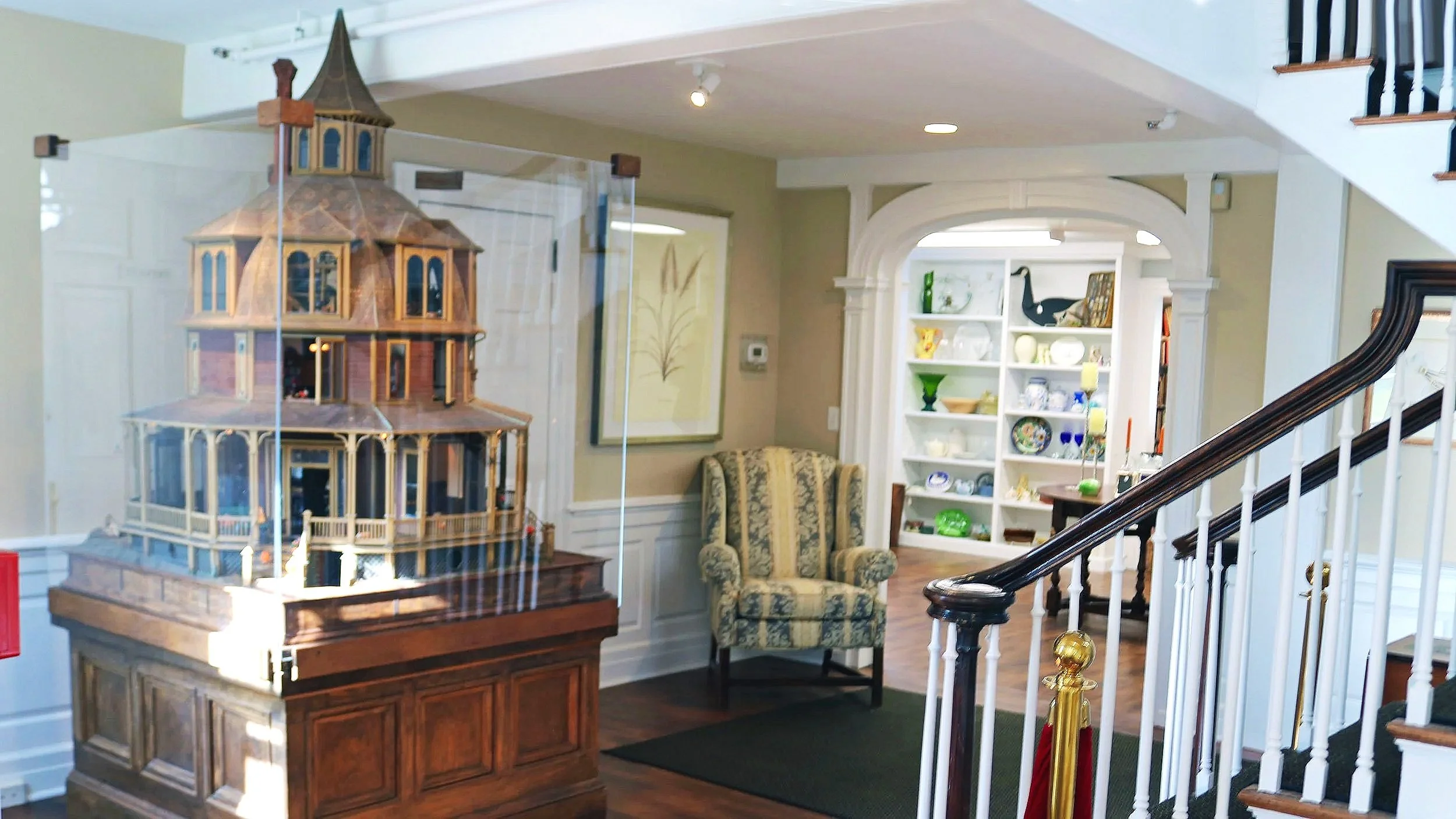 A detailed model of a Victorian-style house inside a glass case on a wooden table in a living room.