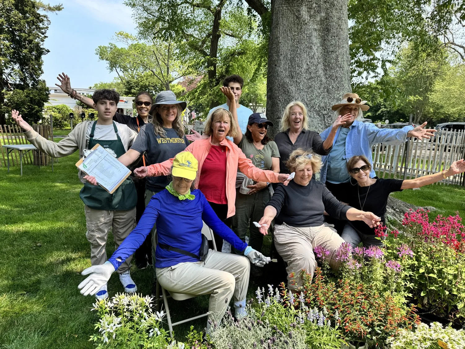 Group of people smiling and posing outdoors near a large tree and colorful flowers, enjoying a sunny day.