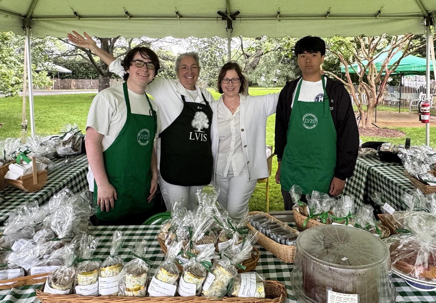 Four people standing behind a table filled with wrapped baked goods at an outdoor fundraiser booth, under a white canopy with trees and green grass in the background.
