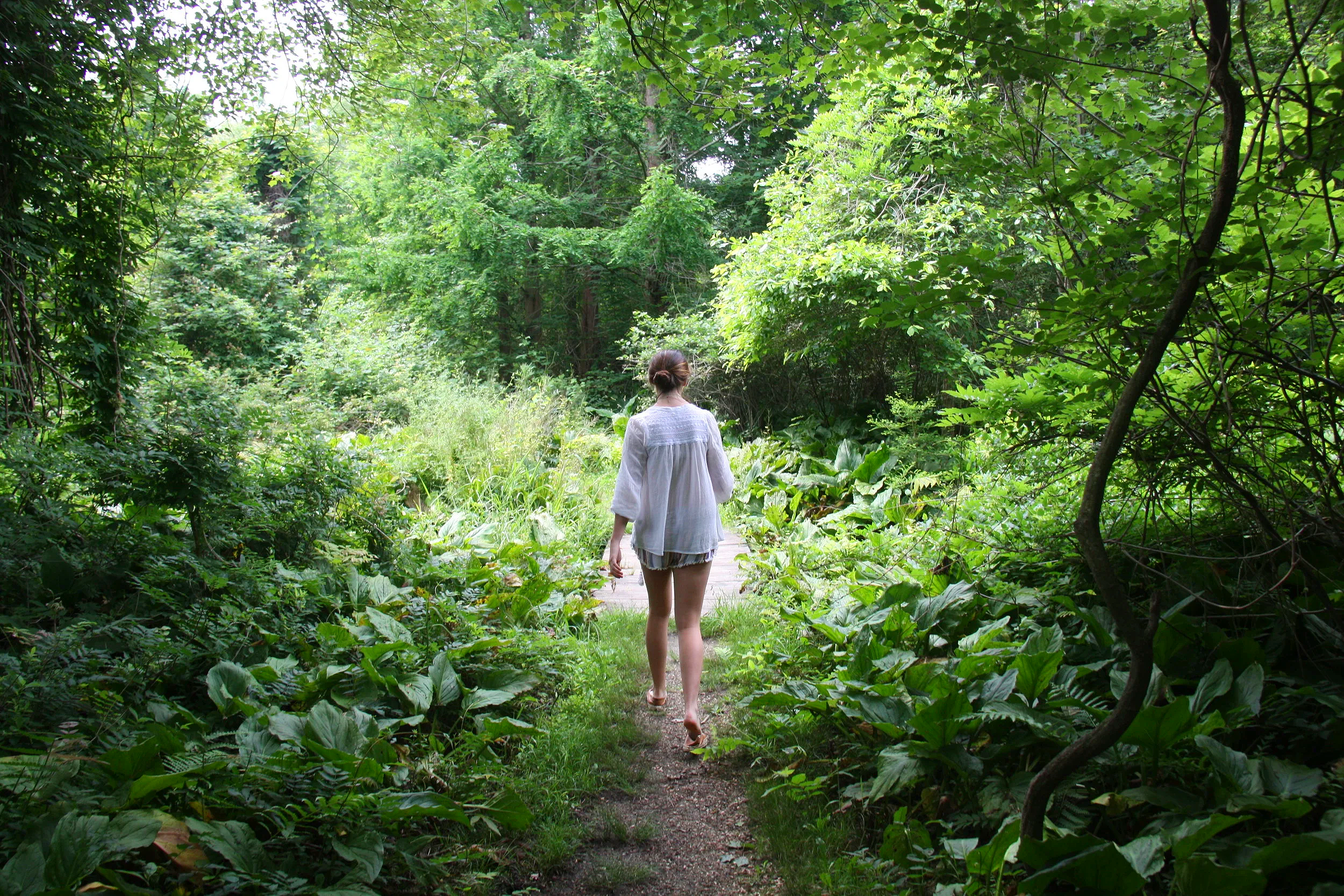 A woman walking down a narrow dirt path through a lush, green forest with dense foliage overhead.