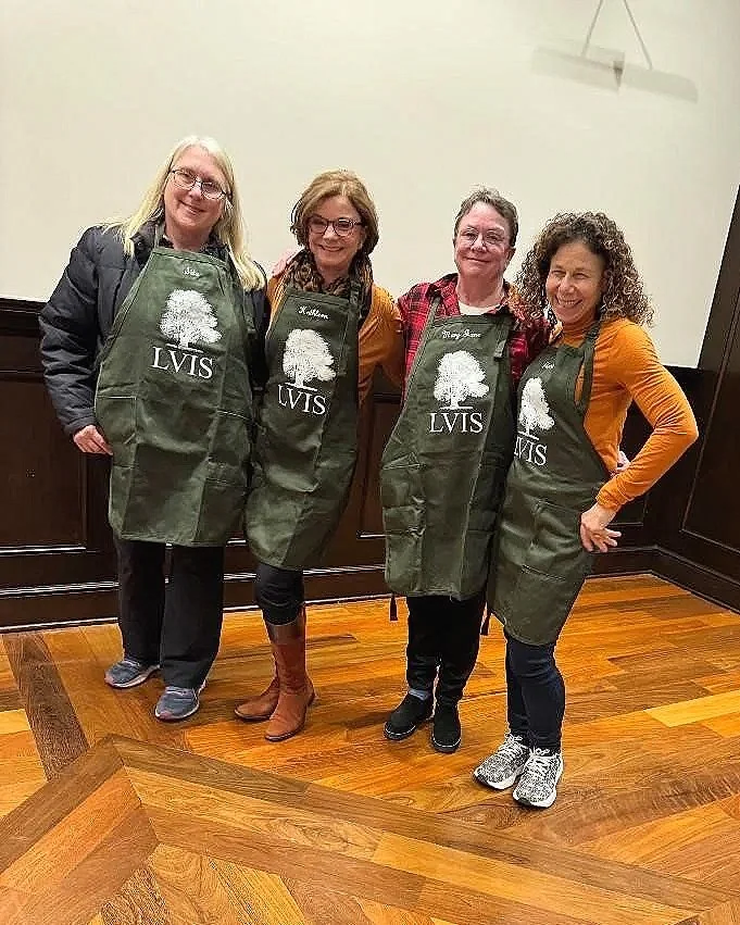 Four women standing together indoors, each wearing a green apron with a tree and 'LVIS' printed on it, smiling and posing for the photo.