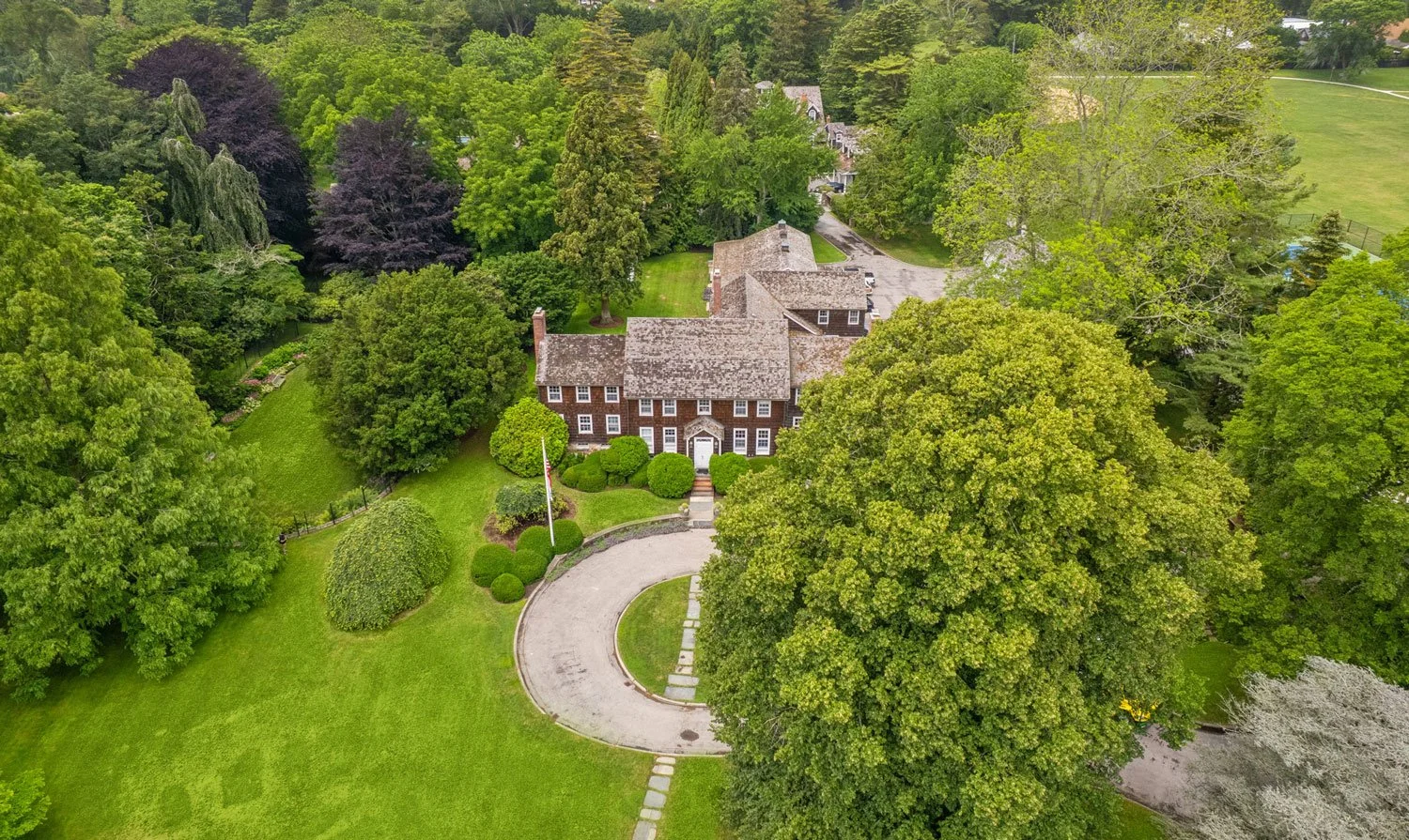 Aerial view of a large brick house surrounded by lush green trees and well-manicured lawn with a curved driveway in front.