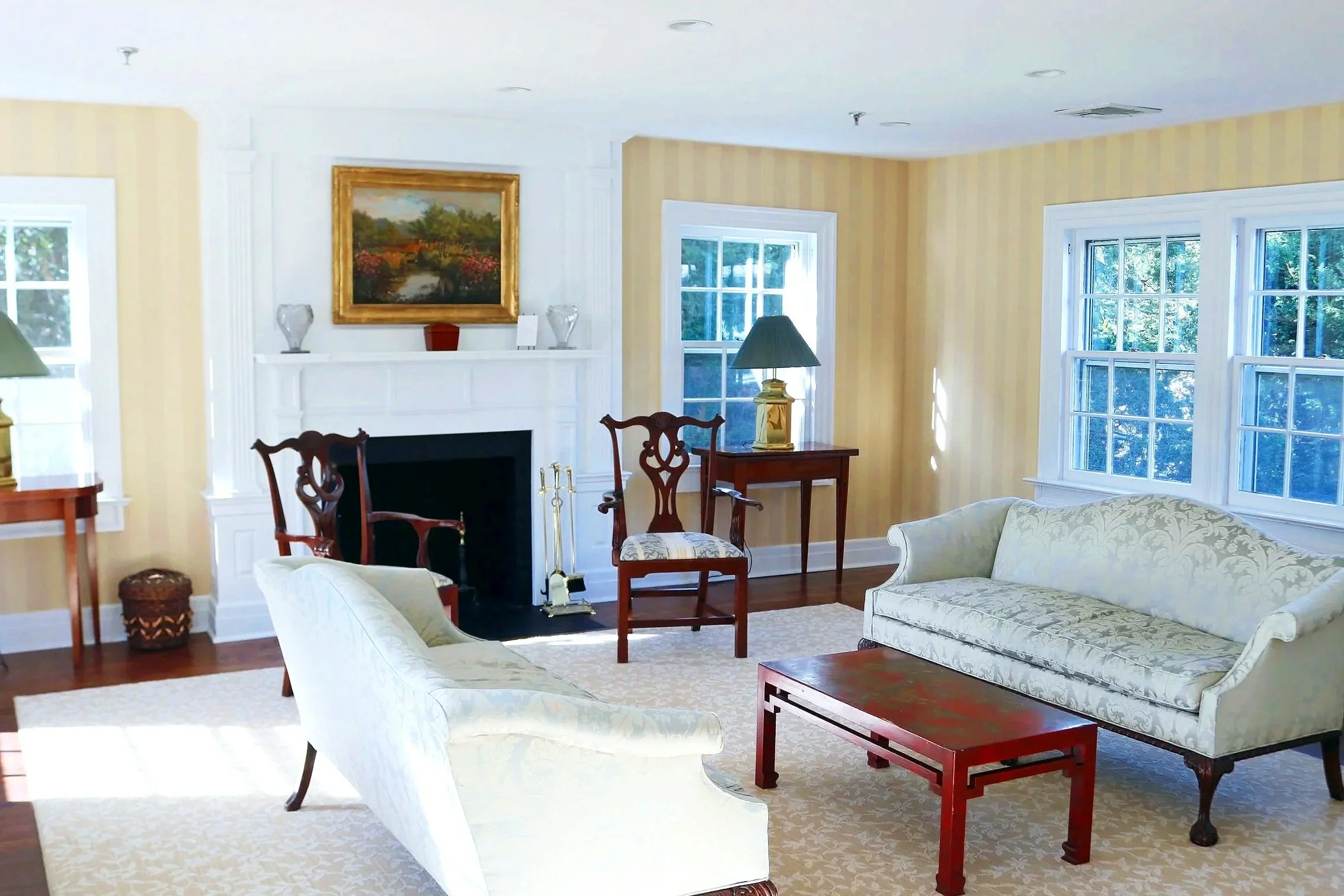 Living room with cream-colored walls, multiple windows, a fireplace with a painting above, and various chairs and tables.