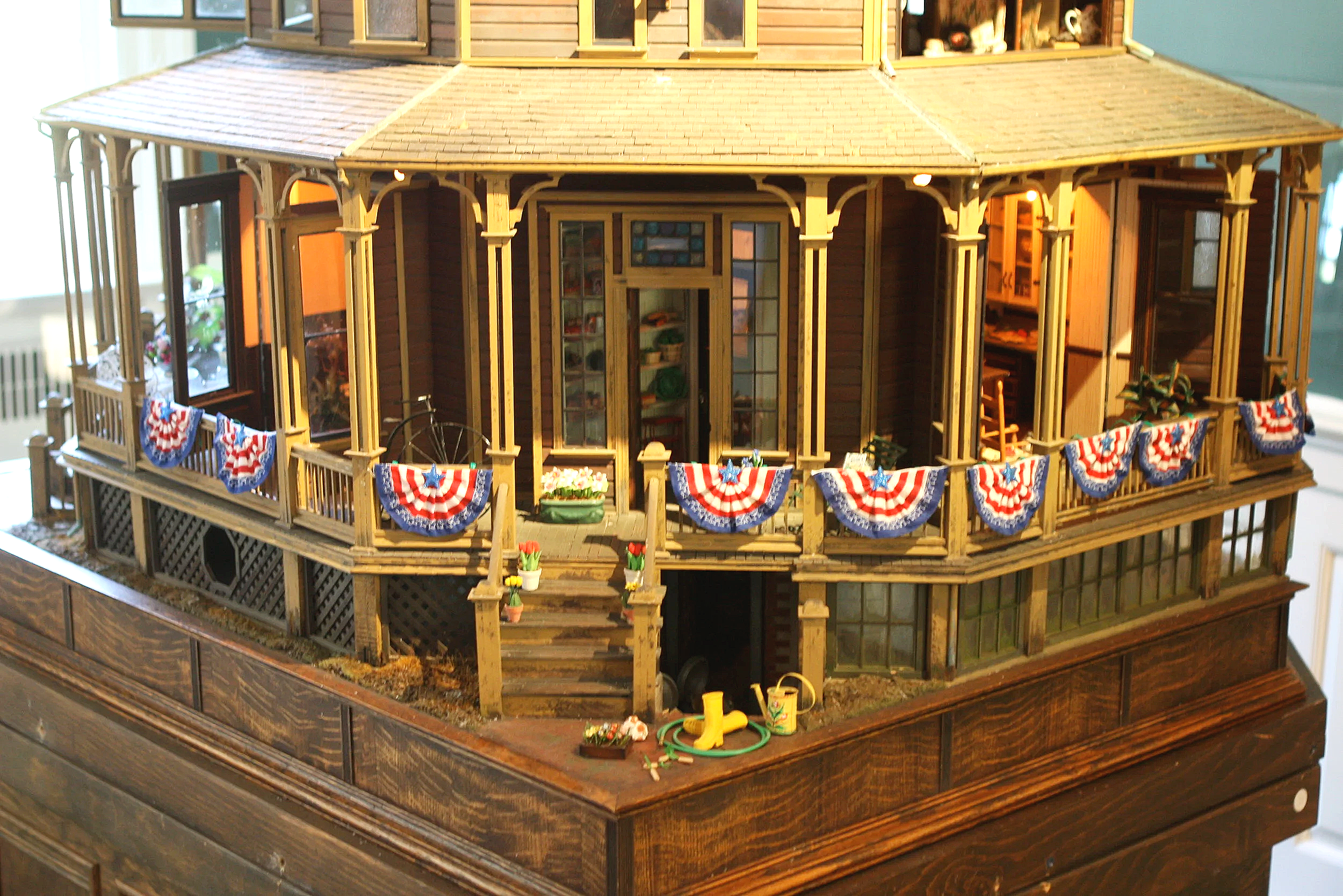 Model of a two-story wooden house with a front porch decorated with red, white, and blue patriotic bunting, lit from inside, with gardening tools and toys on the ground nearby.