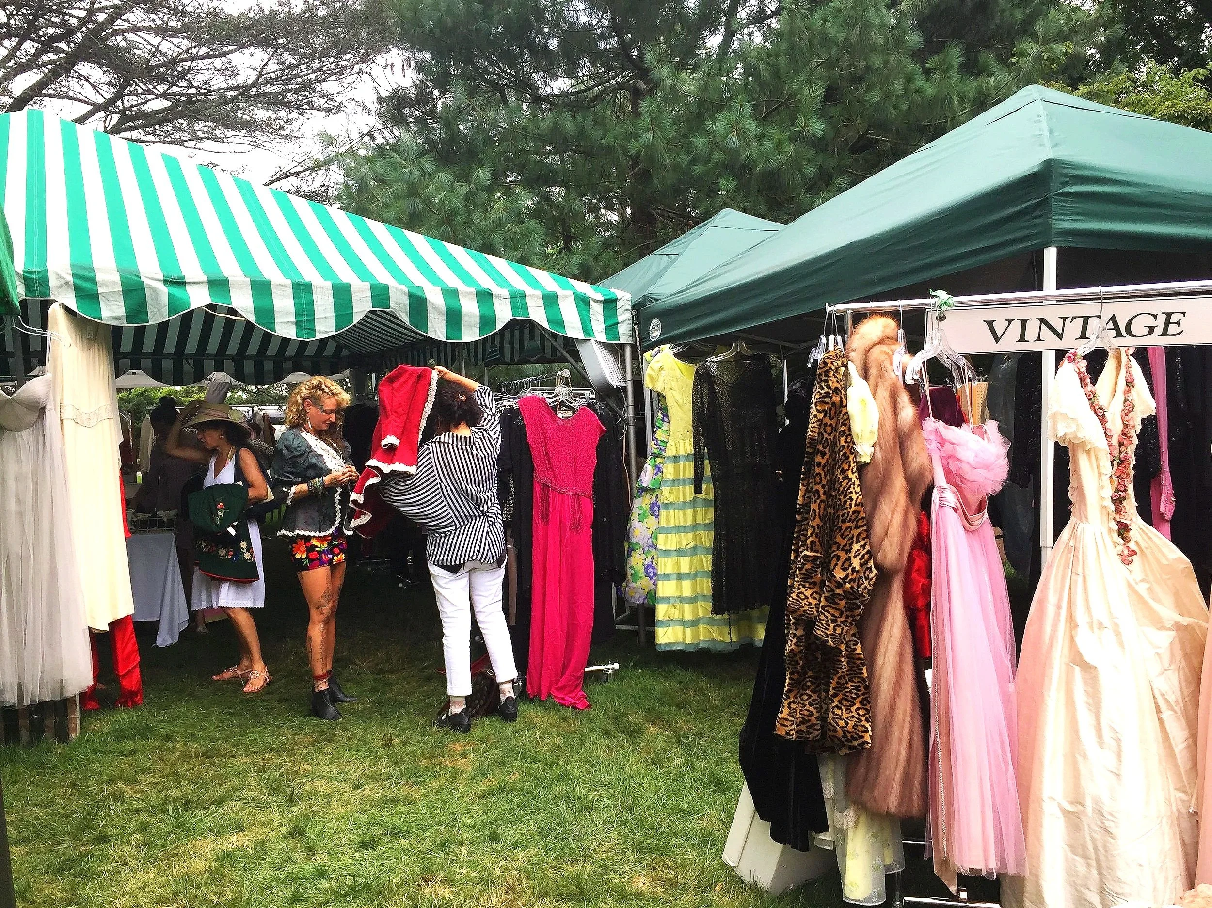 People shopping at an outdoor vintage clothing market under green and white striped tents, with various dresses on display and a group of women examining clothing.