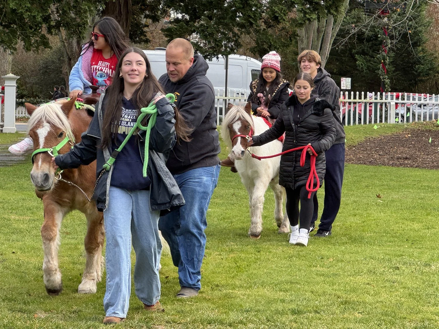 A group of five young people and an adult riding and leading small horses in an outdoor park with a white fence, trees, and grass.