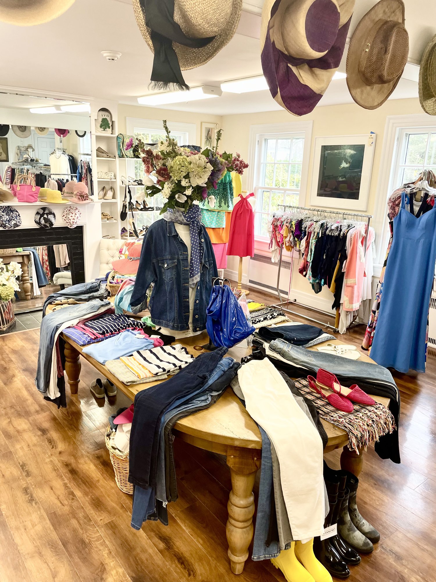Indoor thrift store with clothing, shoes, hats, and accessories displayed on tables, racks, and mannequin. Wooden floors, large windows, and ceiling lights. Hats hanging from ceiling, floral arrangement on mannequin, and various folded clothes on table.