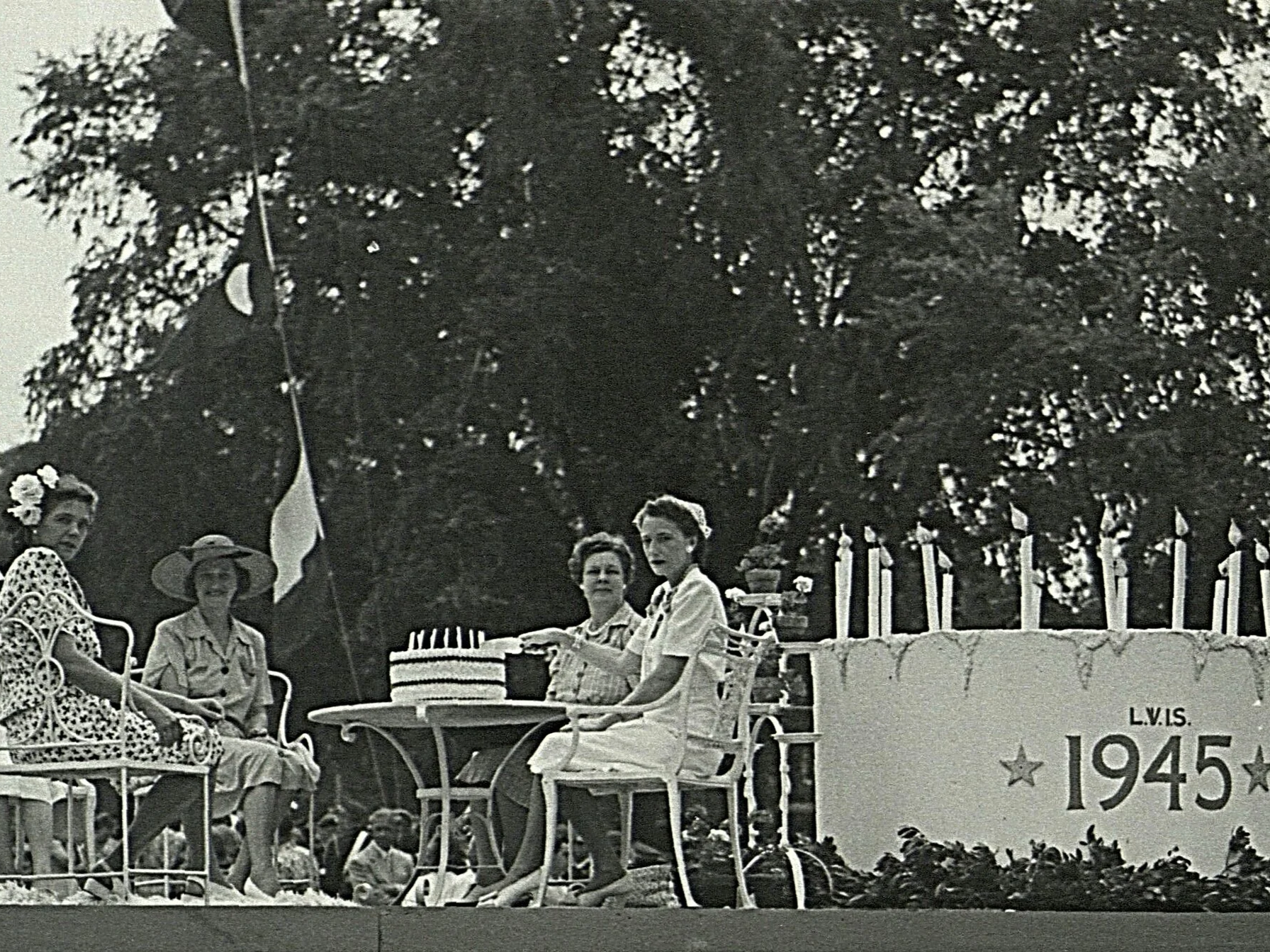 A black and white photo of four women sitting and standing on a decorated float or platform outdoors, possibly celebrating an event from 1945, with trees and flowers in the background.