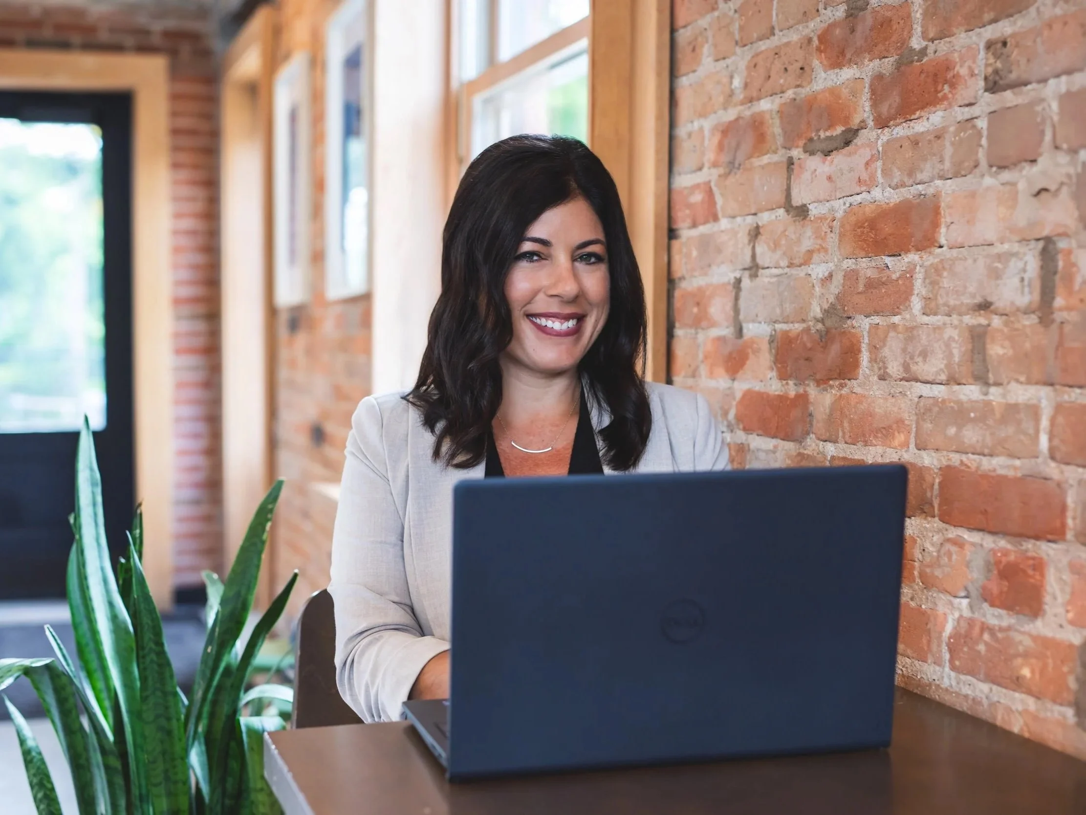 A woman with dark wavy hair smiling and working on a laptop at a desk in a modern office with exposed brick wall, plants, and framed pictures.