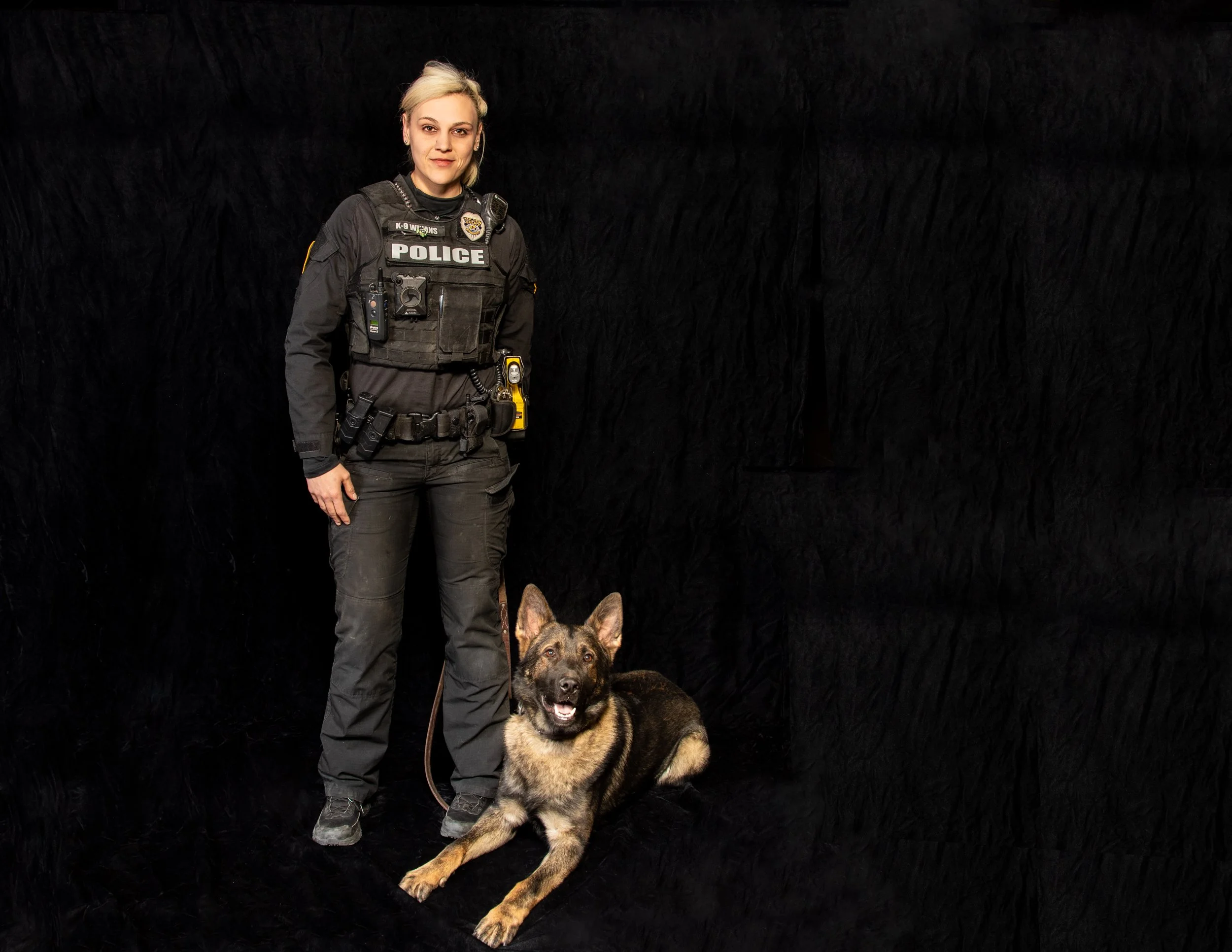 A Tucson Police Department (TPD) officer in full uniform standing next to her seated German Shepherd K9 partner against a black studio backdrop.