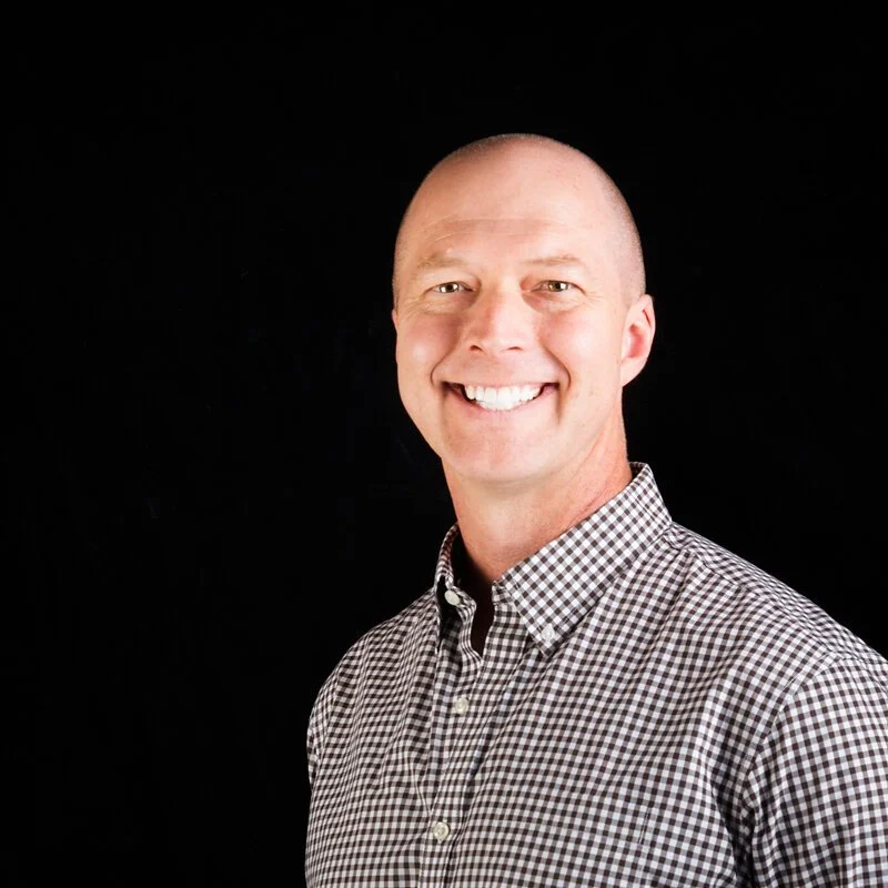 A smiling man with a bald head wearing a checkered button-up shirt, against a solid black background.