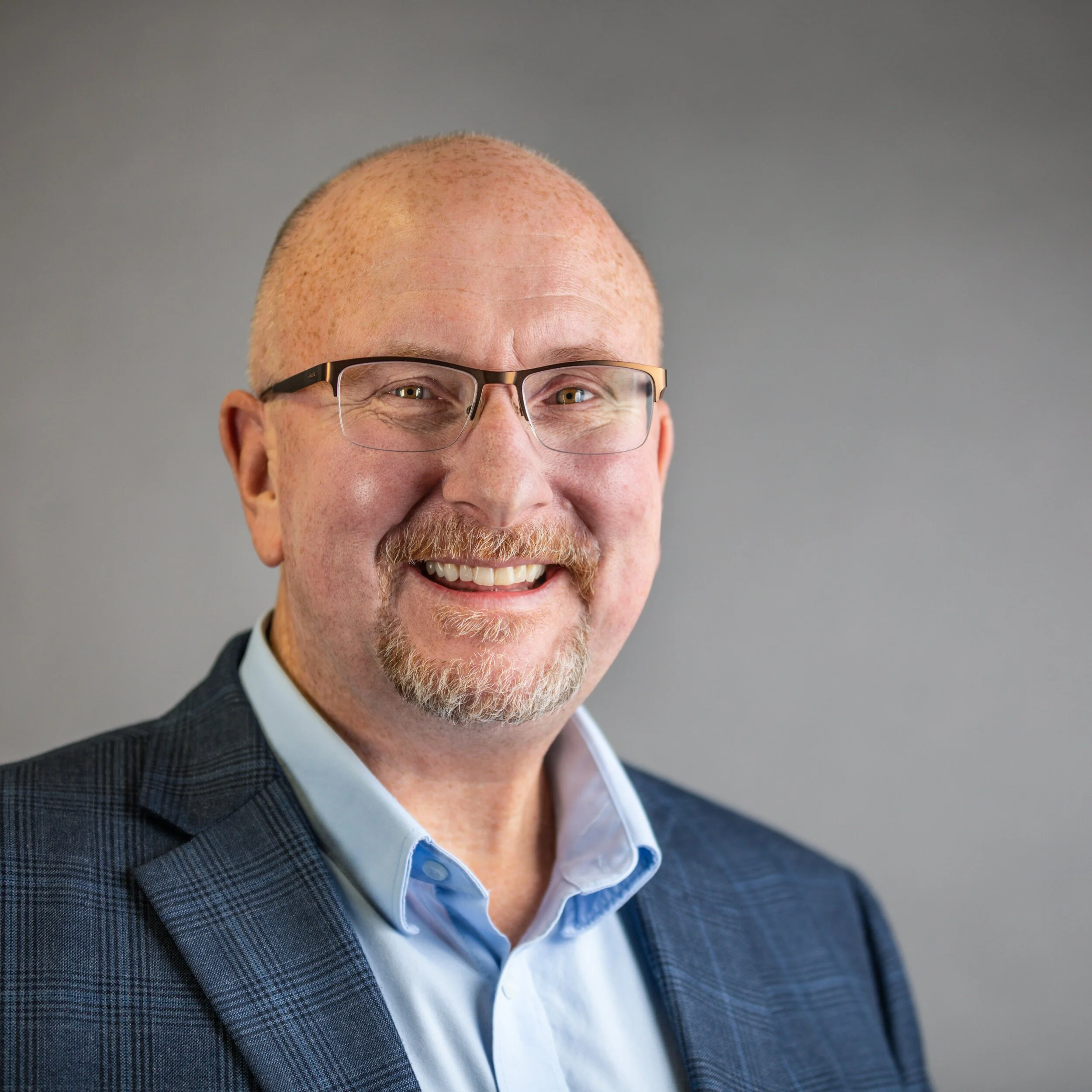Headshot of a smiling man with glasses, a beard, and a bald head, wearing a blue checkered blazer and a light blue collared shirt, against a gray background.