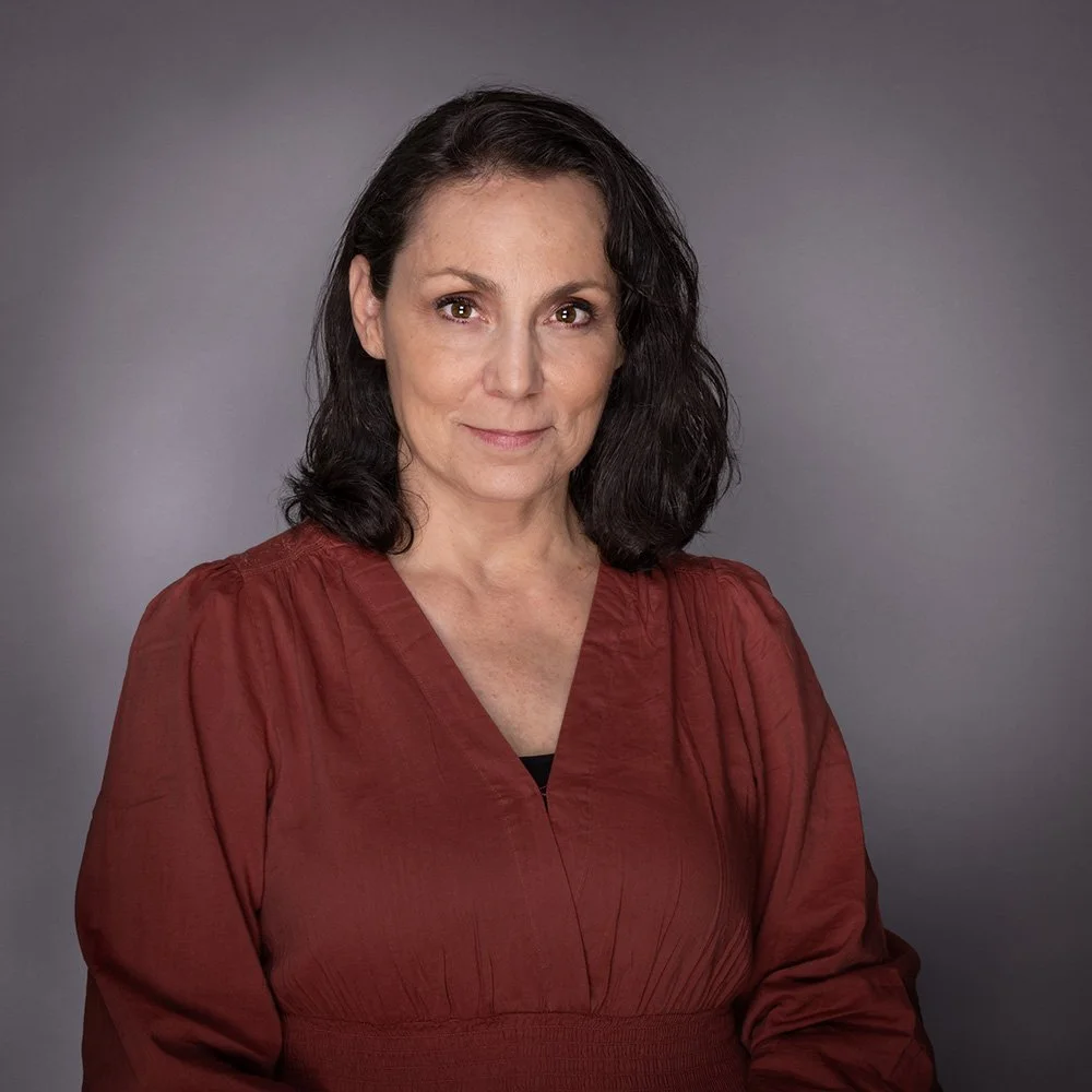 A woman with dark hair wearing a reddish-brown top, posing against a gray background.