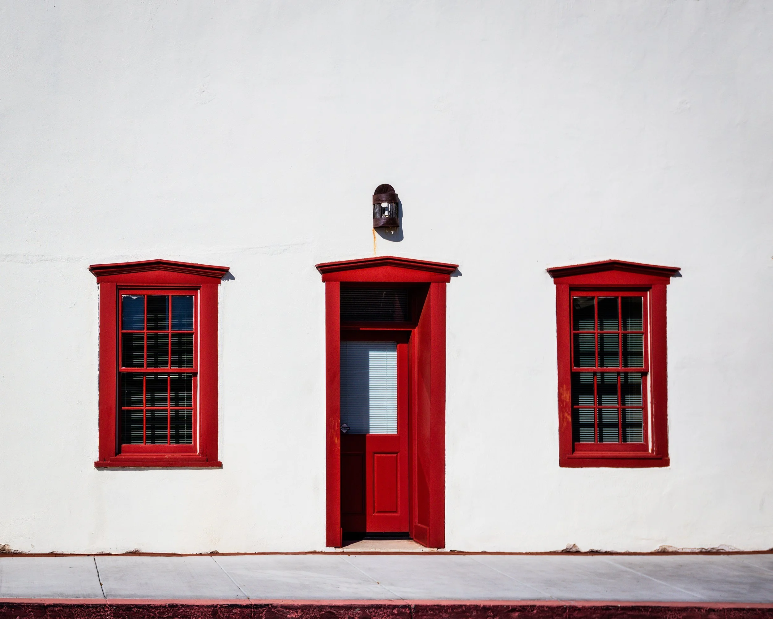 White building with two red window frames and a red door, black light fixture above door, sidewalk in front in the Tucson Museum of Art historic block.