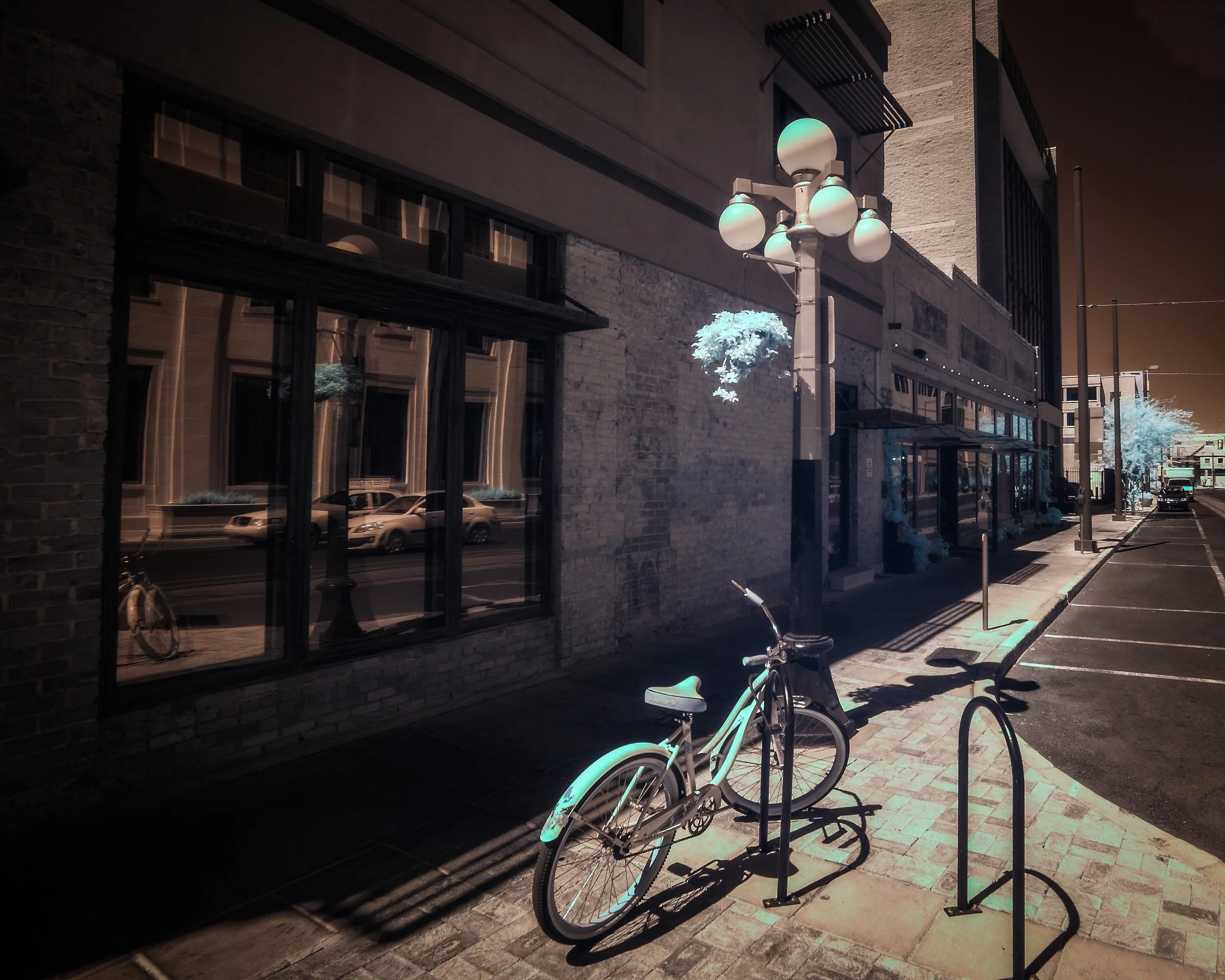 An infrared photograph of a Tucson sidewalk with a bicycle locked to a bike rack, a streetlamp with multiple round lights, and building with large windows reflecting parked cars and trees with illuminated foliage.
