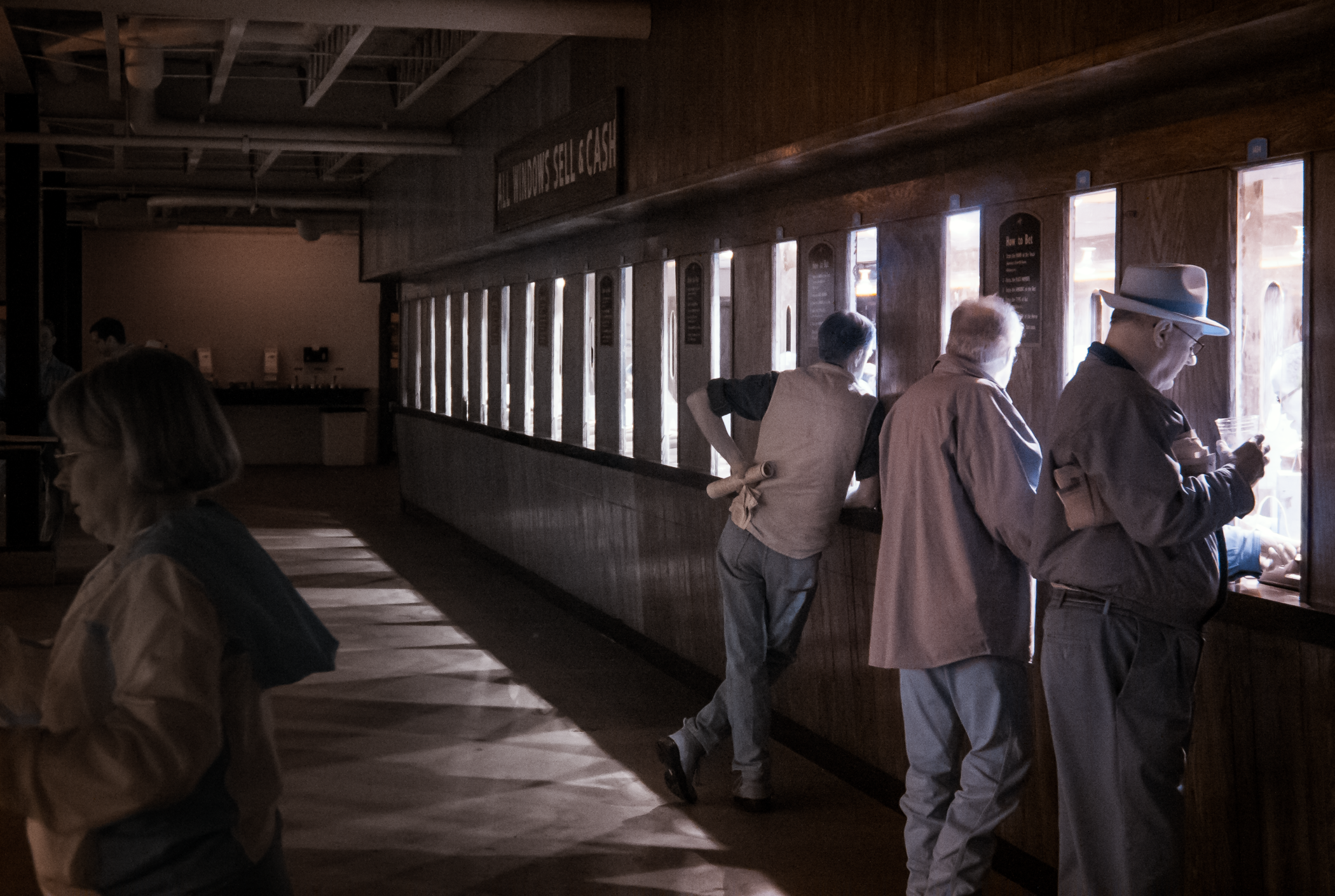 People standing at a betting counter windows of a wooden interior, at Keeneland Race Track in Lexington, KY, with a woman with glasses in the foreground and three older men wearing coats and hats at the windows.