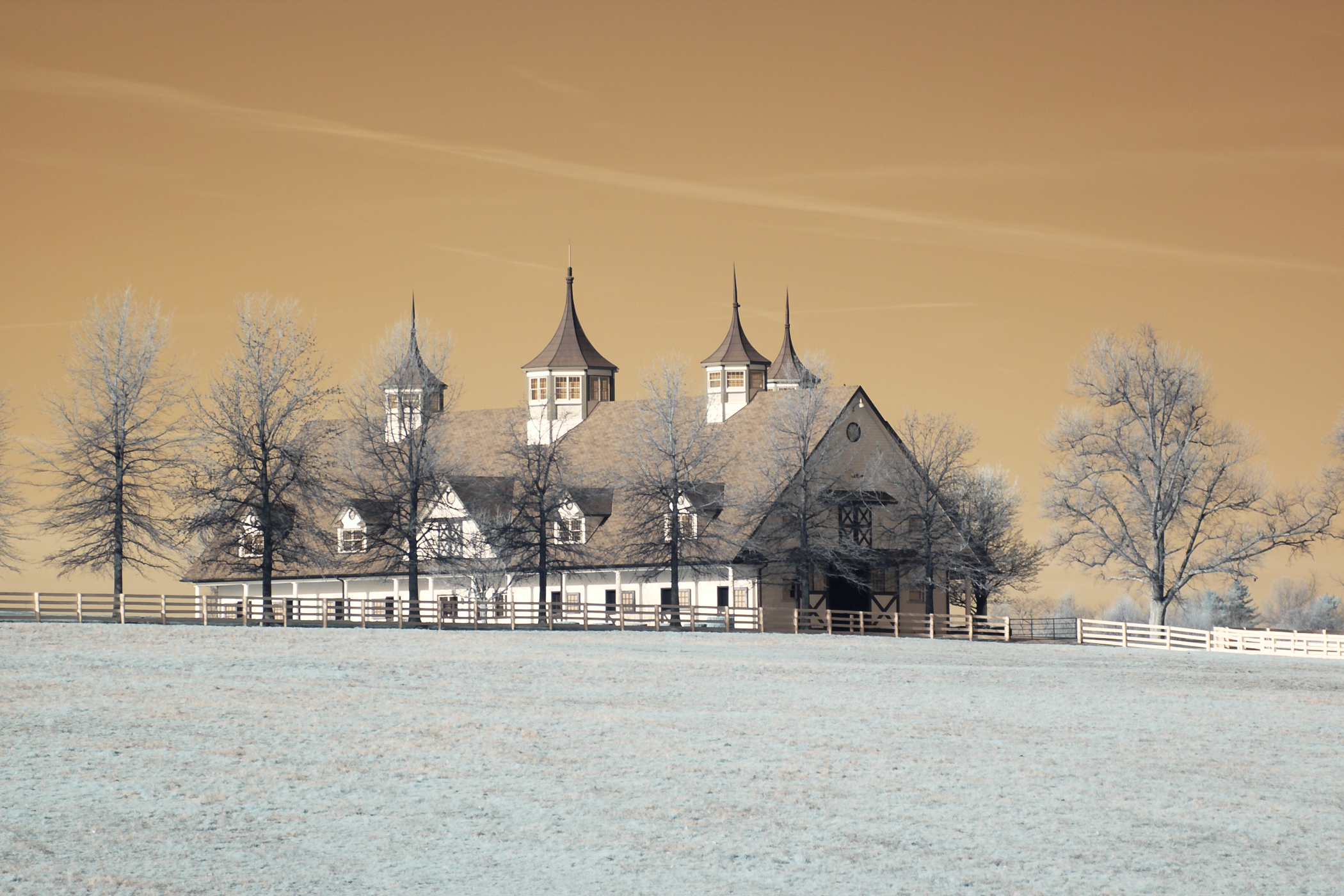 An old-fashioned barn with multiple towers and steeples, surrounded by leafless trees and a wooden fence, under a yellowish sky.