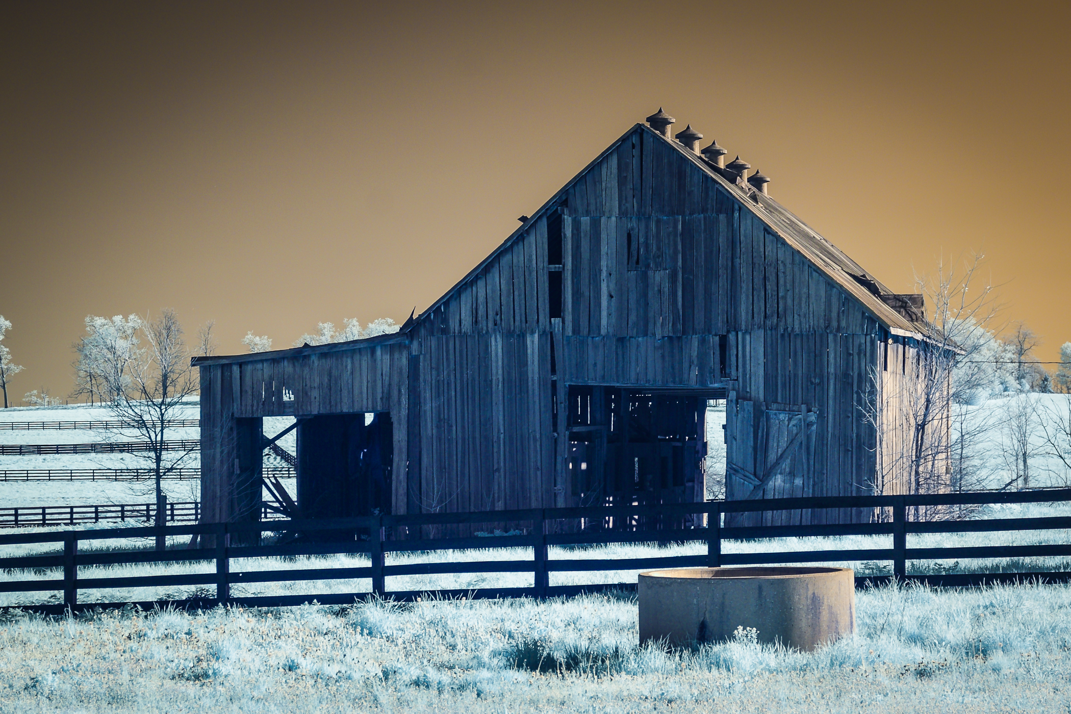 An old weathered wooden barn in a snowy field with a black fence, leafless trees in the background, and a cloudy sky.