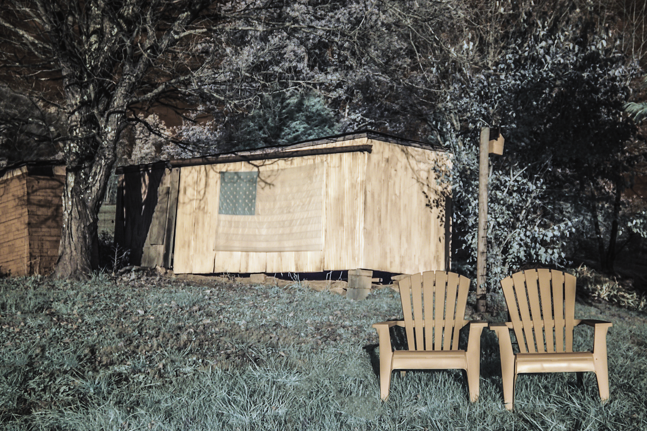 Nighttime backyard scene with two beige Adirondack chairs on grass, a wooden shed with an American flag window, and trees in the background.