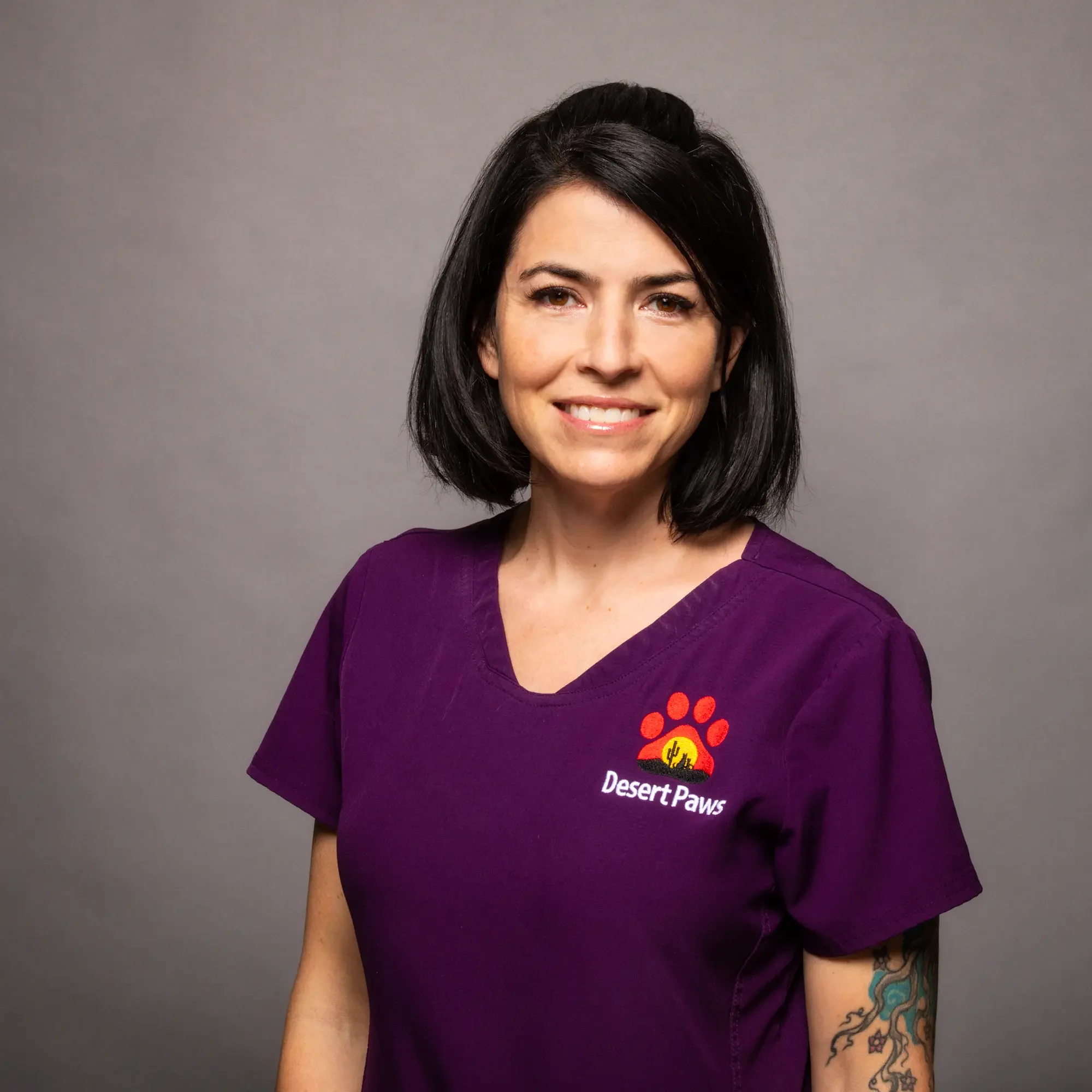 Portrait of a smiling woman with black hair, wearing a purple Desert Paws scrub top, standing against a gray background.