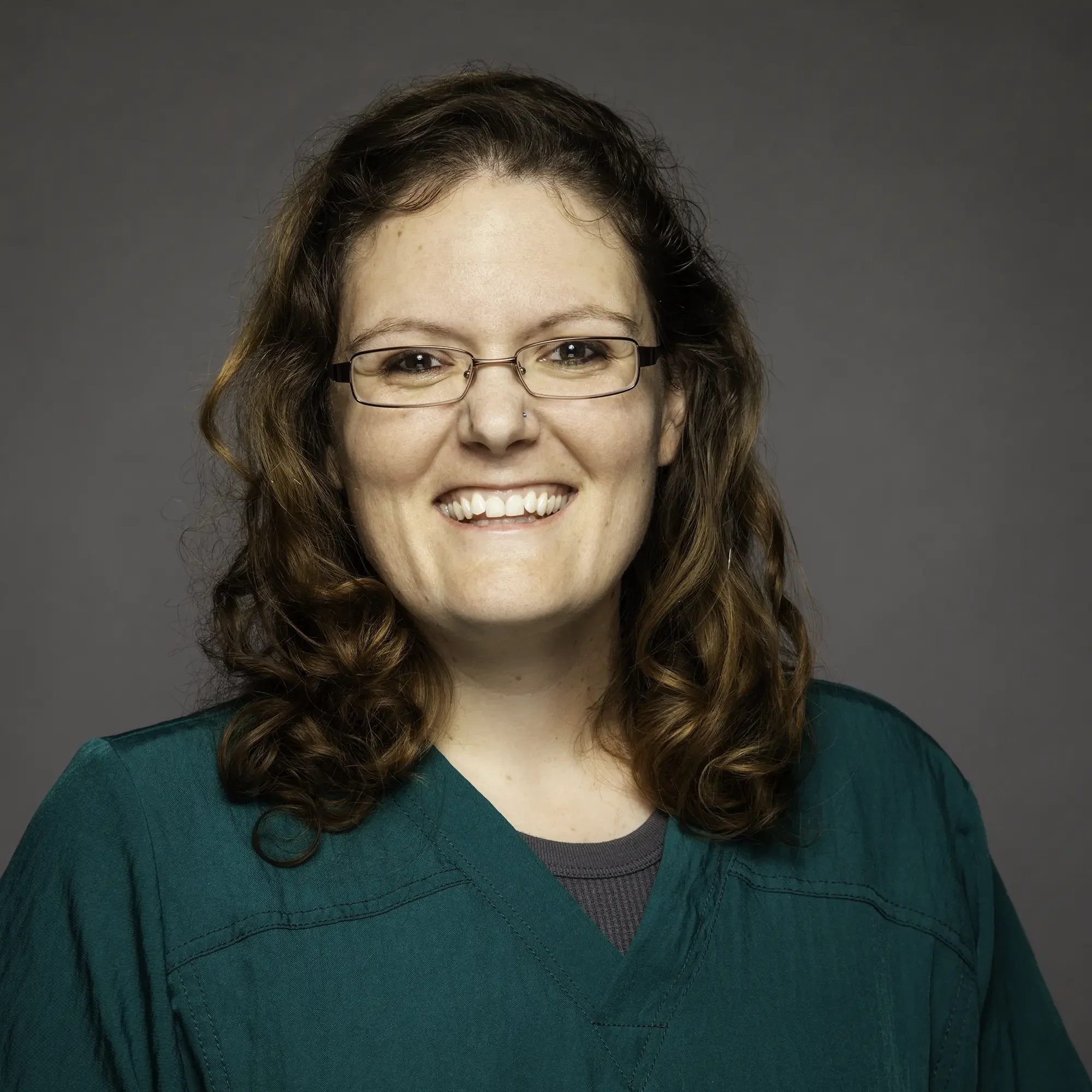 A woman with curly brown hair, glasses, and a nose piercing, wearing a teal top, smiling against a gray background.