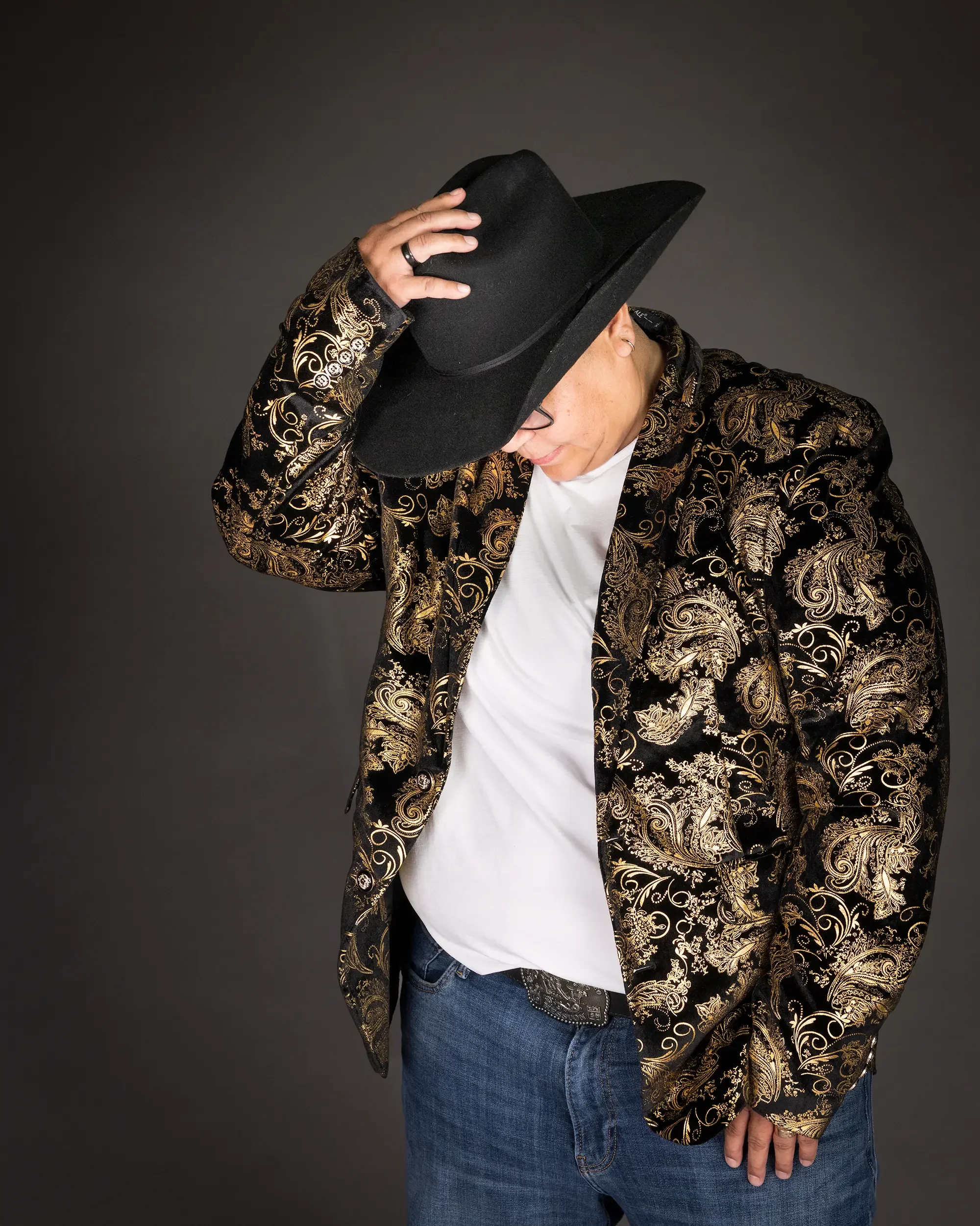 A portrait of a woman putting on a cowboy had wearing a shiny gold jacket and a white t-shirt in front of a gray background.