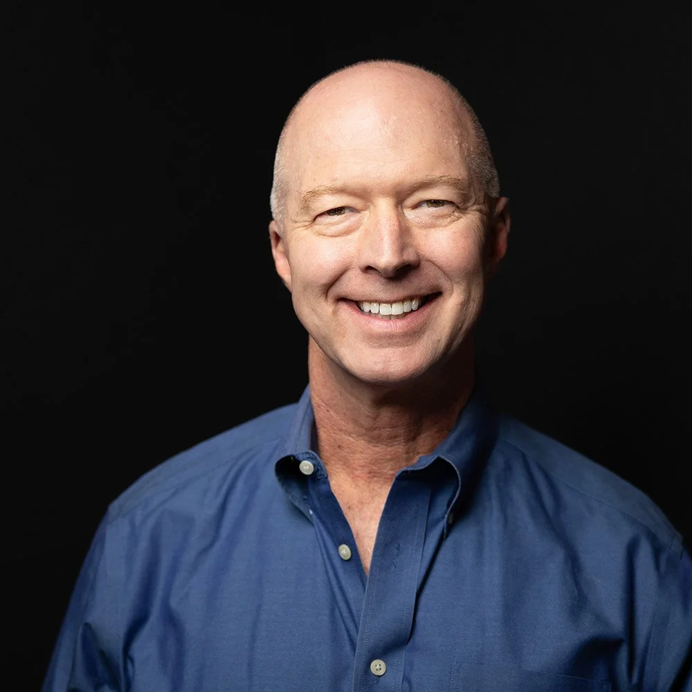 A smiling middle-aged man with a bald head, wearing a blue button-up shirt, against a black background.