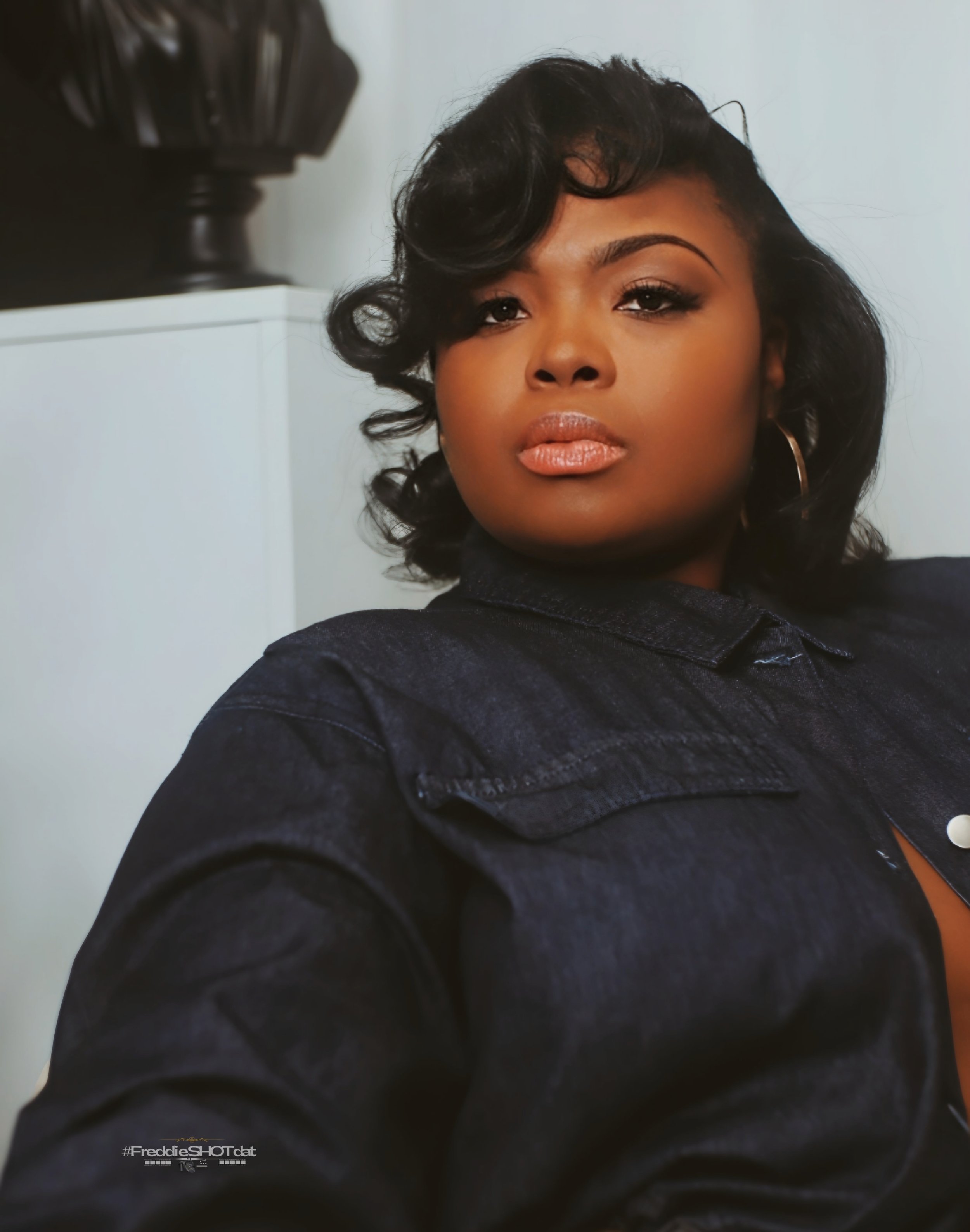 Close-up of a woman with short, curly black hair, wearing makeup, hoop earrings, and a black denim jacket, looking confidently at the camera.