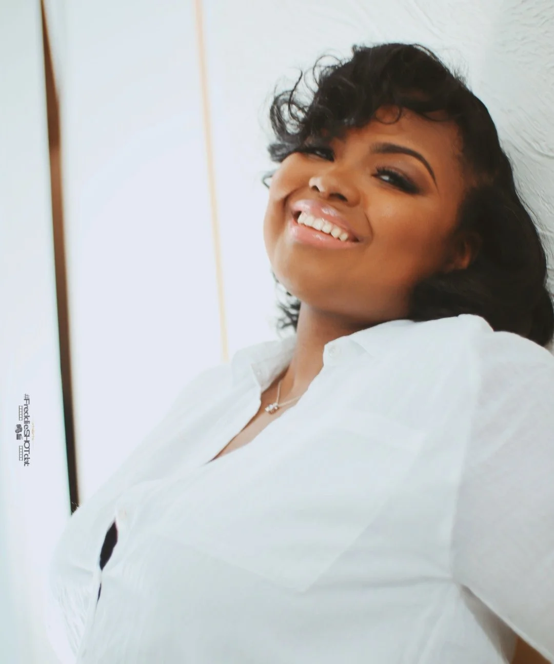 A woman with dark curly hair smiling and tilting her head back, wearing a white shirt and standing against a white wall.