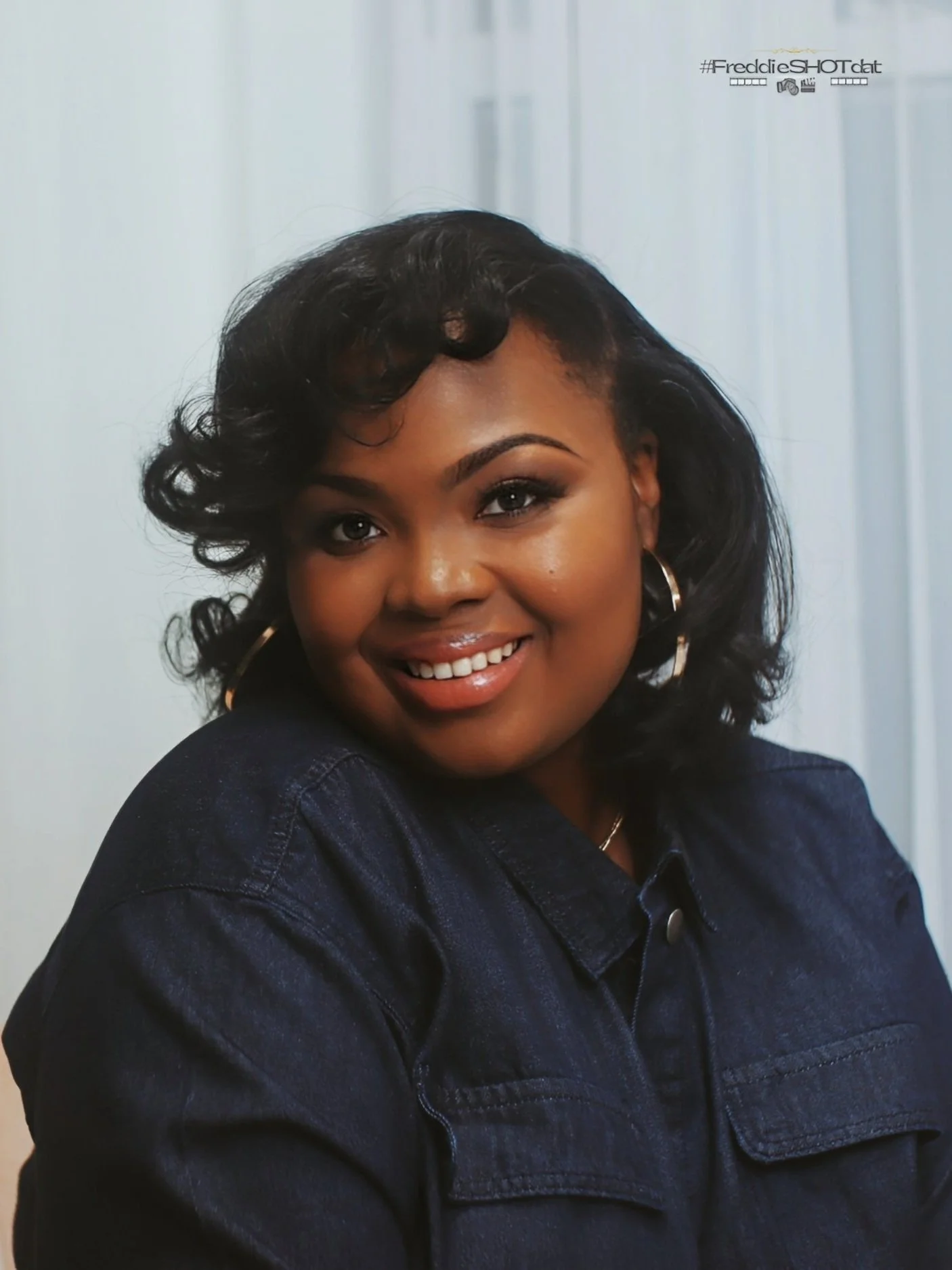 A smiling woman with dark, curly hair wearing hoop earrings and a dark denim jacket portrait.