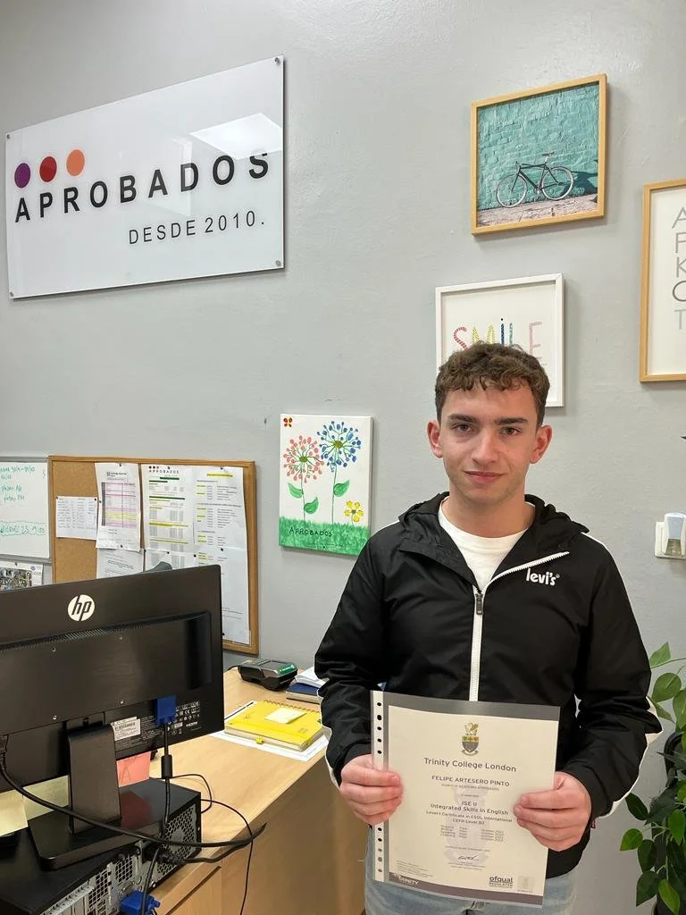 Joven sosteniendo un diploma de Trinity College London en una oficina decorada con pinturas y carteles motivacionales.
