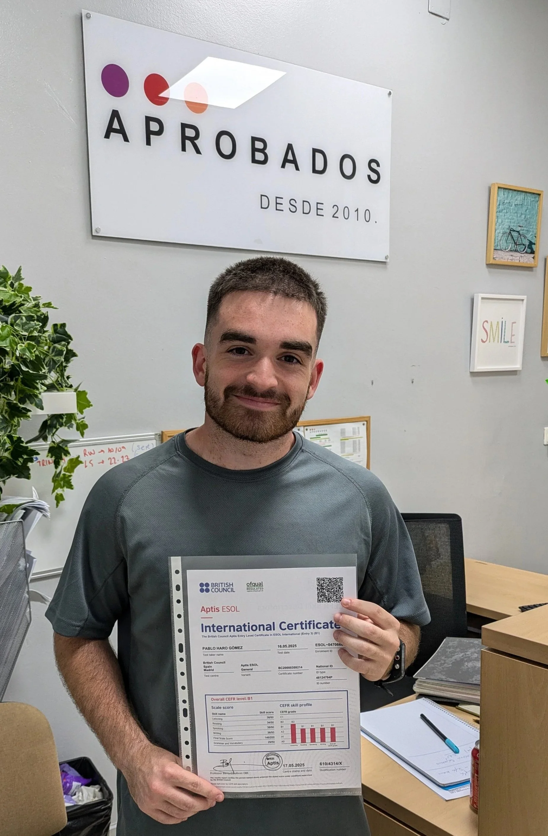 Hombre sosteniendo un certificado de B1 en la academia, con cartel que dice "Aprobados desde 2010" en la pared tras él.