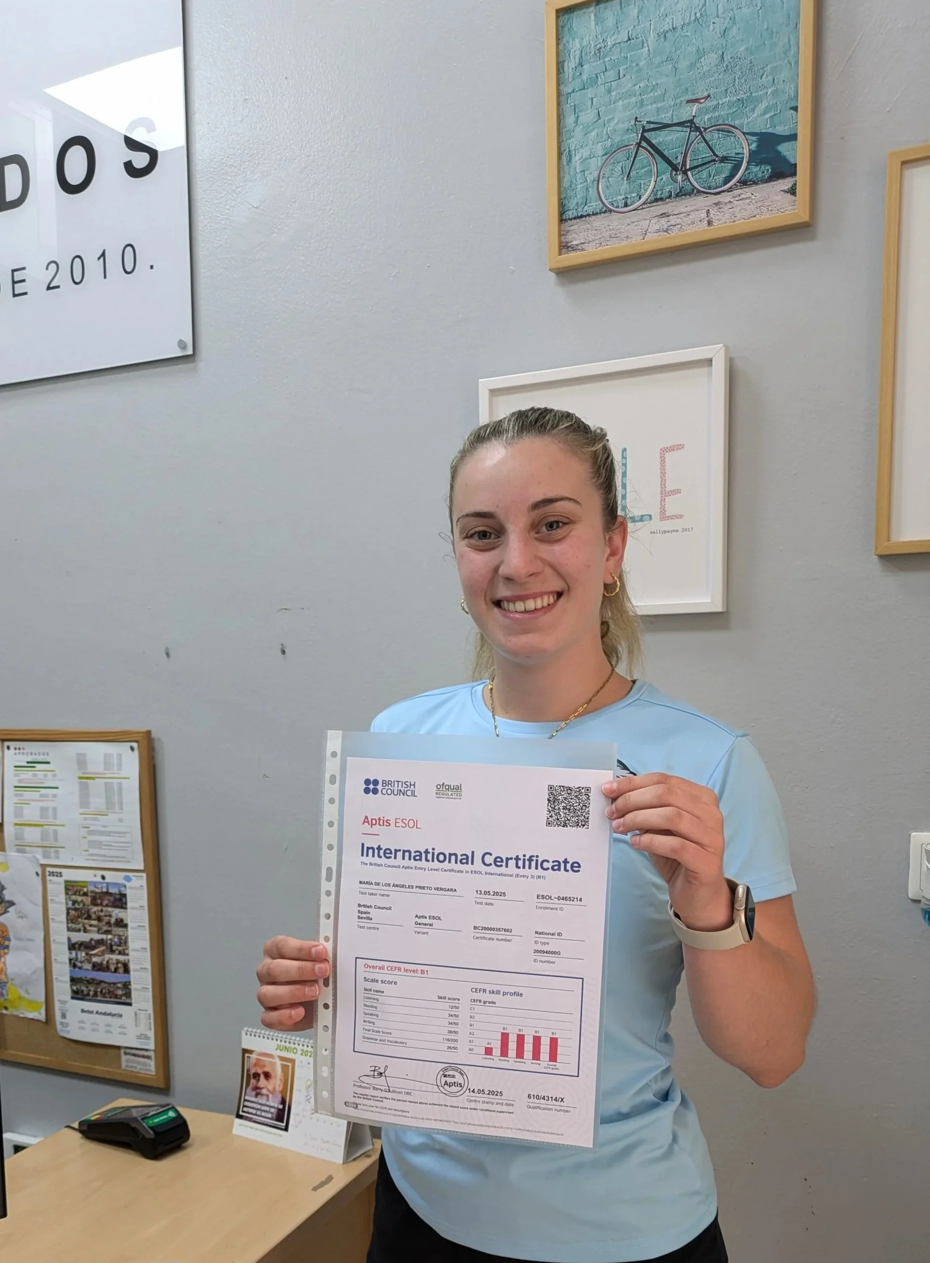 Mujer sonriendo y sosteniendo un certificado frente a una pared decorada con cuadros y anuncios de la Academia.