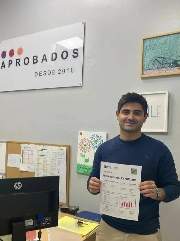 Hombre sonriendo sosteniendo un certificado en la academia con cartel que dice 'Aprobados desde 2010' en la pared.