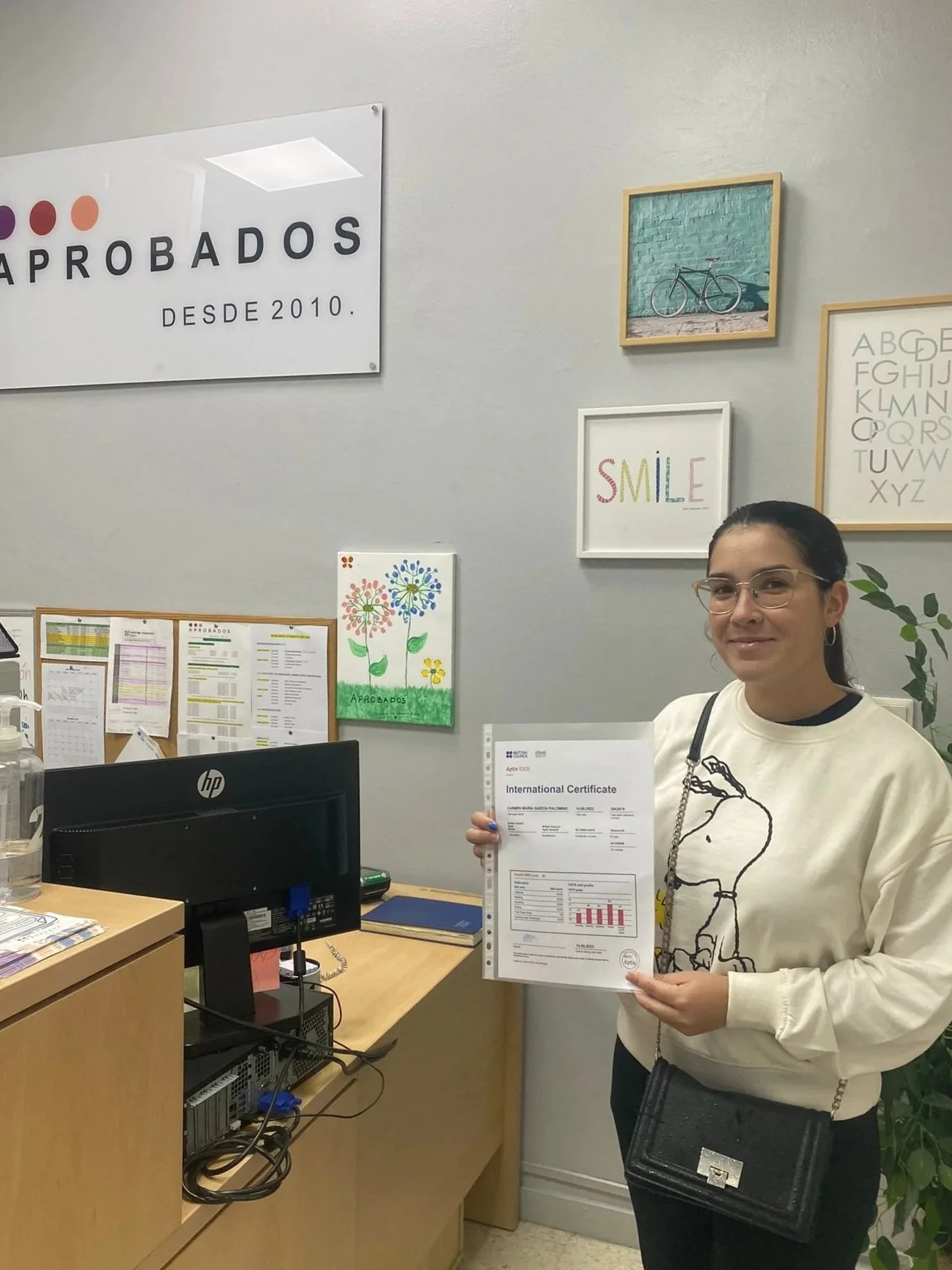 Mujer en oficina mostrando certificado de B1 junto a un ordenador y papeles en escritorio, decoraciones en la pared.