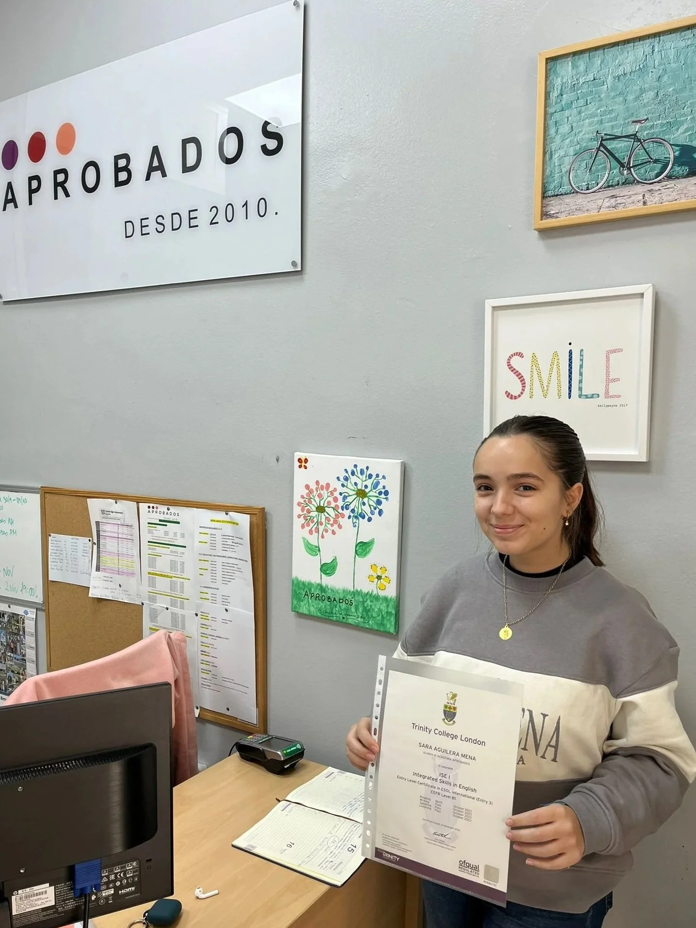 Una joven sostiene un certificado académico frente a un escritorio con ordenador y libros en la recepción de la academia con paredes decoradas con cuadros y carteles, incluyendo uno que dice "Aprobados" desde 2010 y otro que dice "Smile".