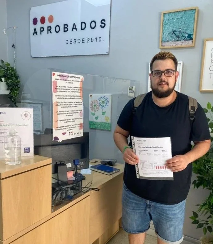 Hombre con gafas y barba, sosteniendo un certificado de reconocimiento, de pie en una oficina con carteles y decoración en la pared, frente a una mesa con un cartel y productos de higiene.