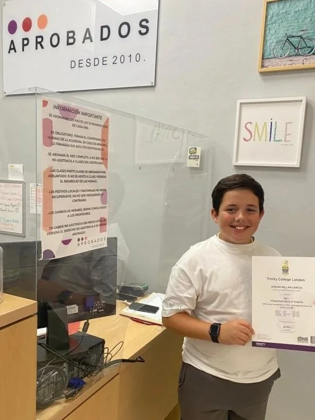 Niño sonriendo sosteniendo un certificado en un aula de un colegio llamado Trinity College London. Comentarios y señalética sobre aprobados en el fondo.